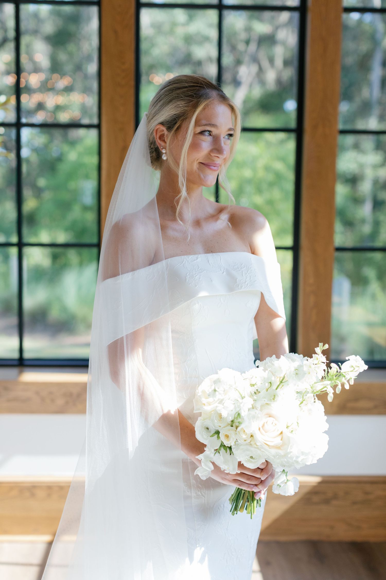 A bride in a white wedding dress holds a bouquet of white flowers while posing near sunlit windows.