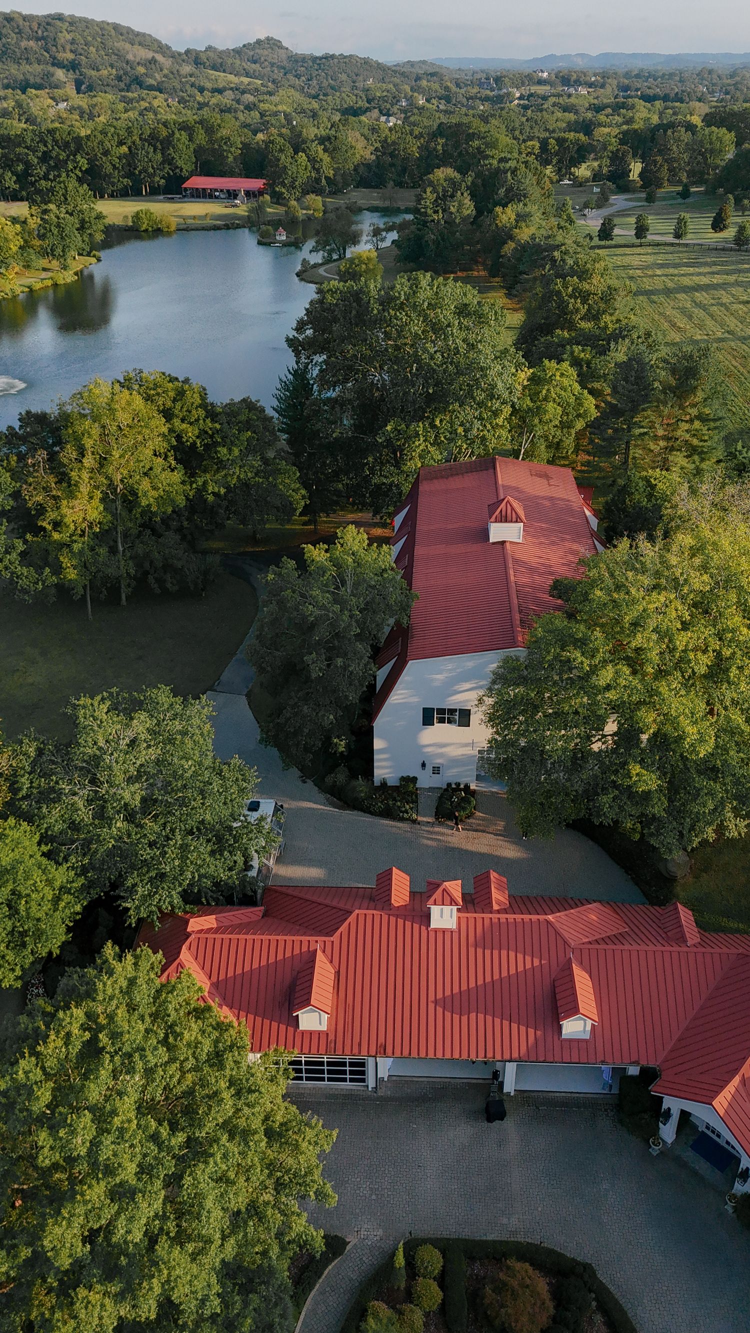 Aerial view of a sprawling ranch-style building complex with red metal roofs surrounded by lush green trees and a lake.