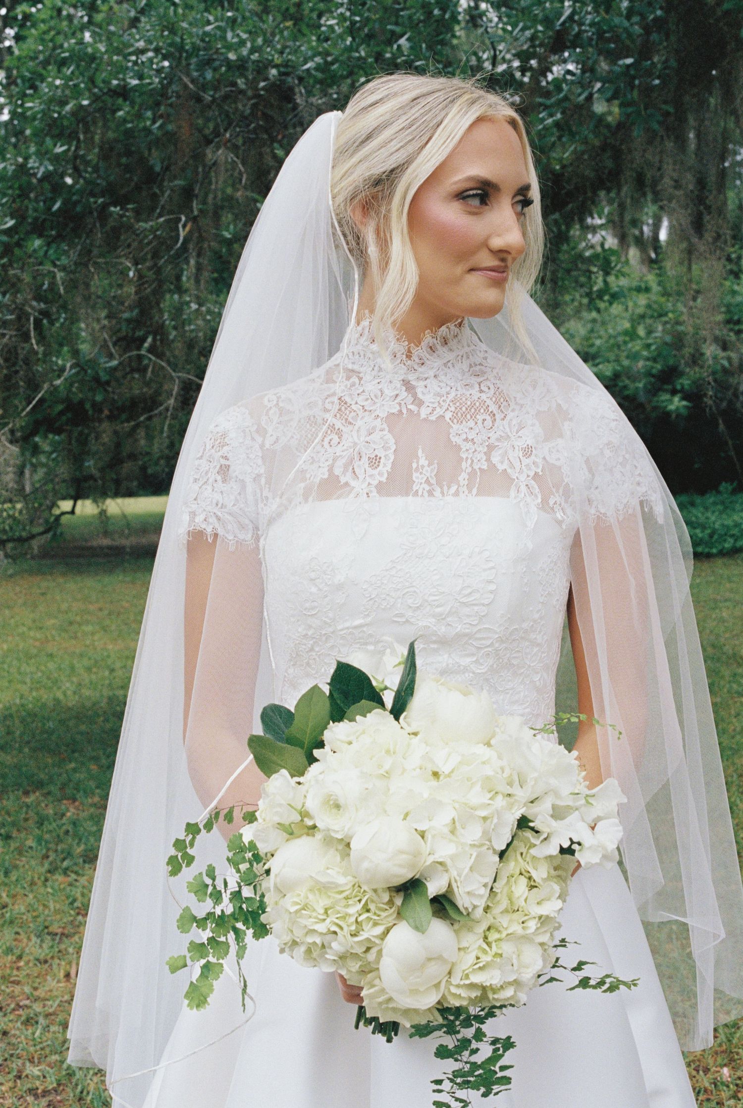 Elegant blonde bride wearing lace high-neck wedding gown and veil holds white floral bouquet outdoors.