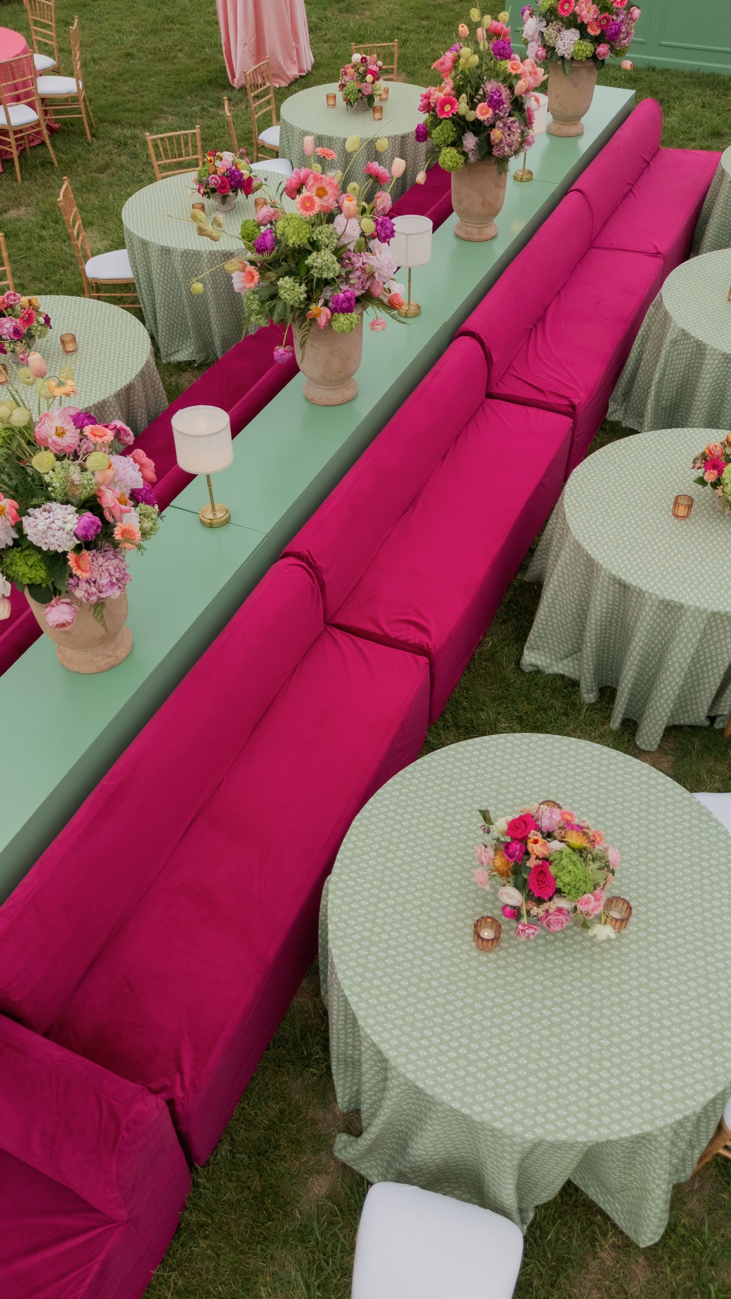 Outdoor event setup with hot pink sofas, mint green tables, and colorful floral centerpieces on lush grass.