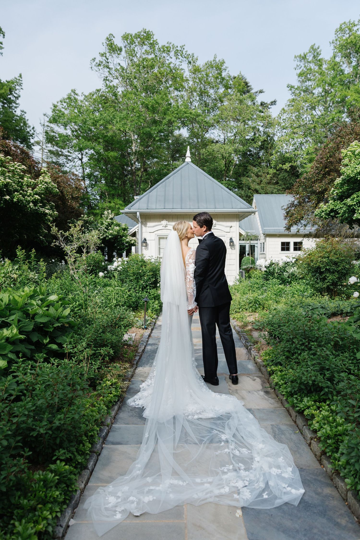 A couple walking down a garden pathway surrounded by greenery with the bride's dress train flowing behind.