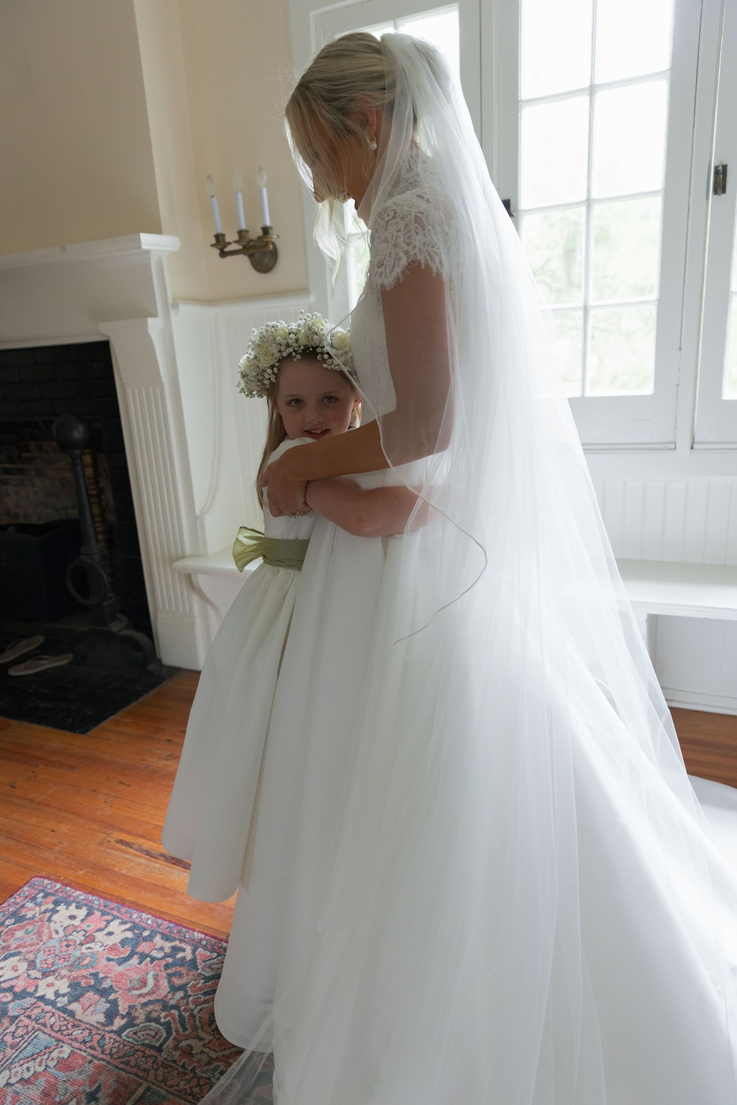 A bride in a white lace veil tenderly embraces a young flower girl holding a bouquet indoors.