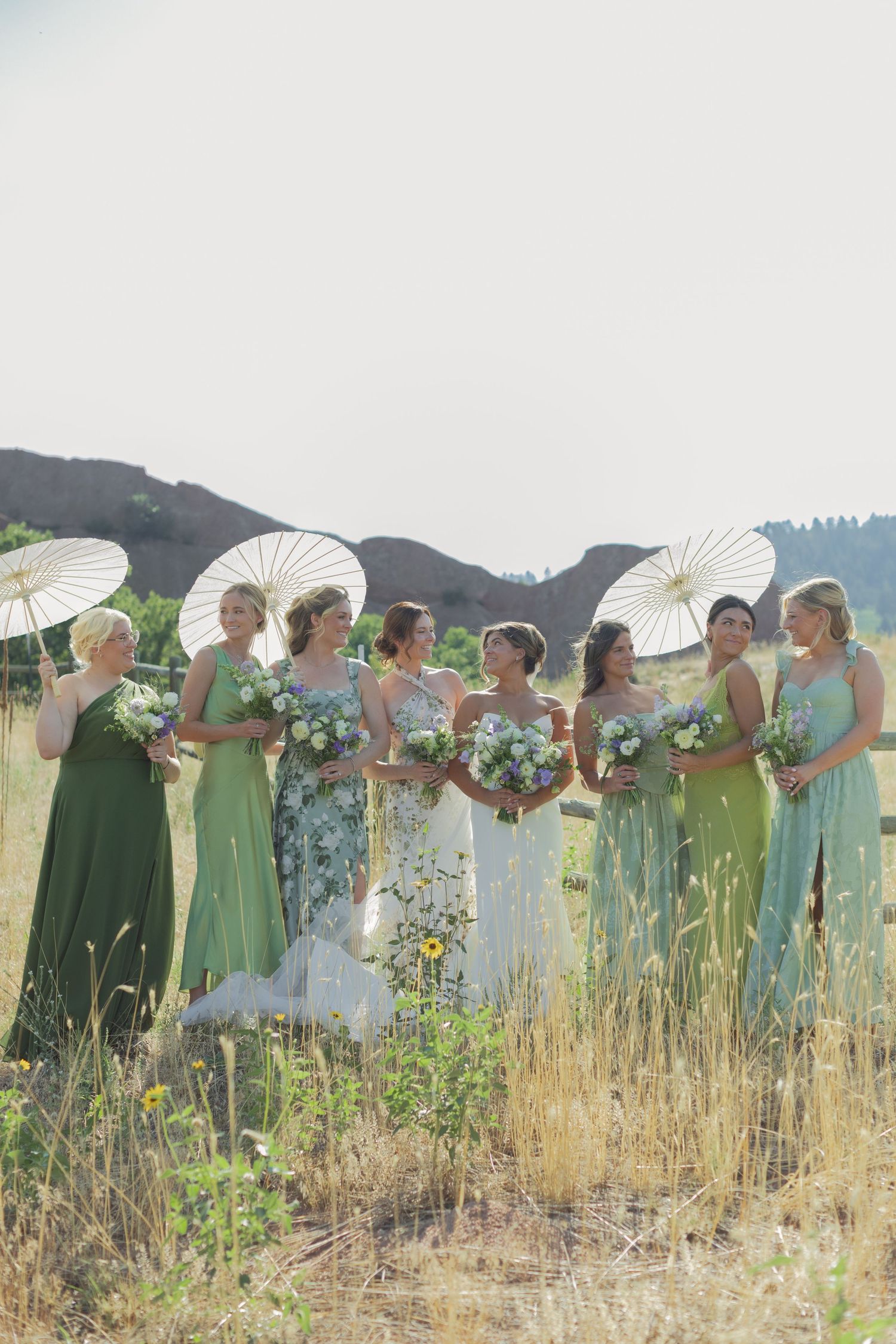 Bride and bridesmaids in mixed green dresses hold bouquets and white parasols in a sunny grassy field with rocky hills behind.