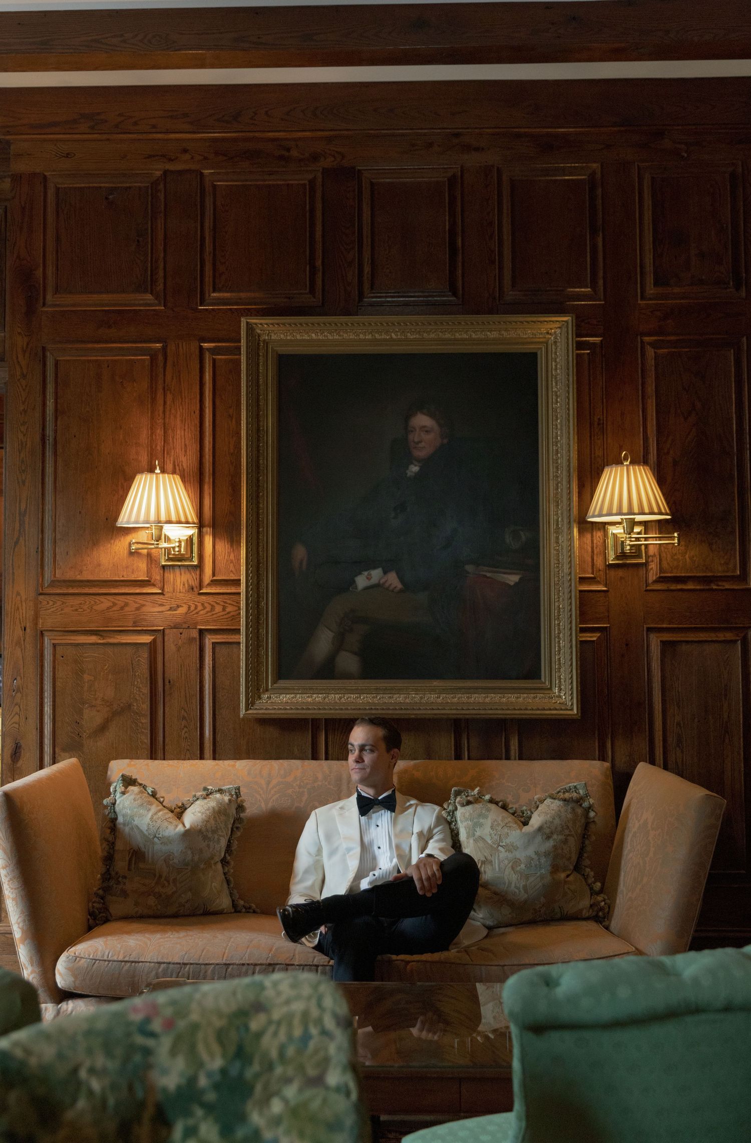 Groom in a white dinner jacket and black bow tie sits on a tan sofa beneath a framed portrait in a wood-paneled room.