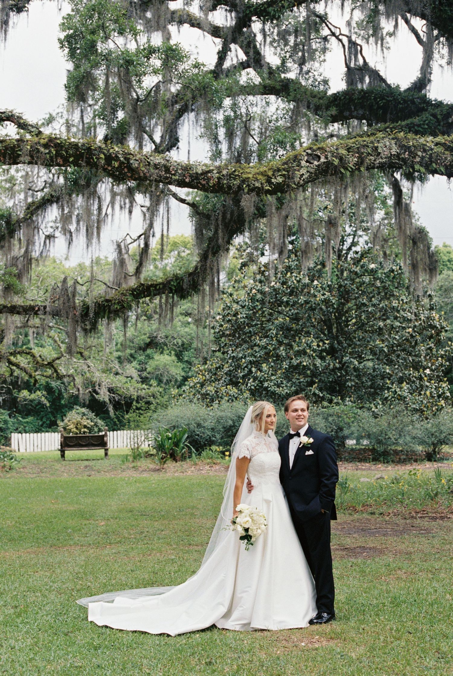 Bride and groom pose under moss-draped oak trees in a lush garden, bride in elegant white gown with veil and train.