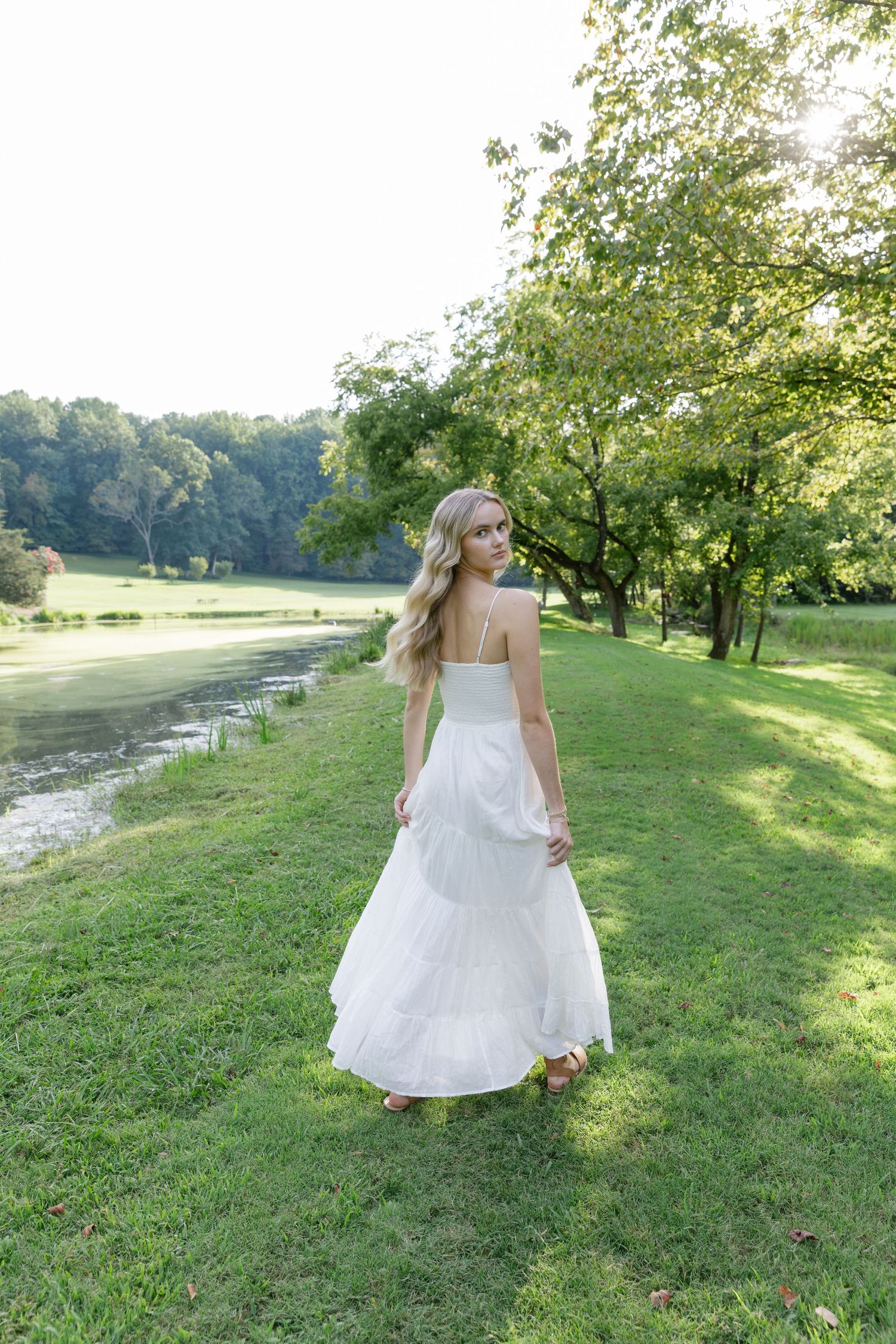 Young woman in white maxi dress standing by a serene pond in a lush green park on a sunny day.
