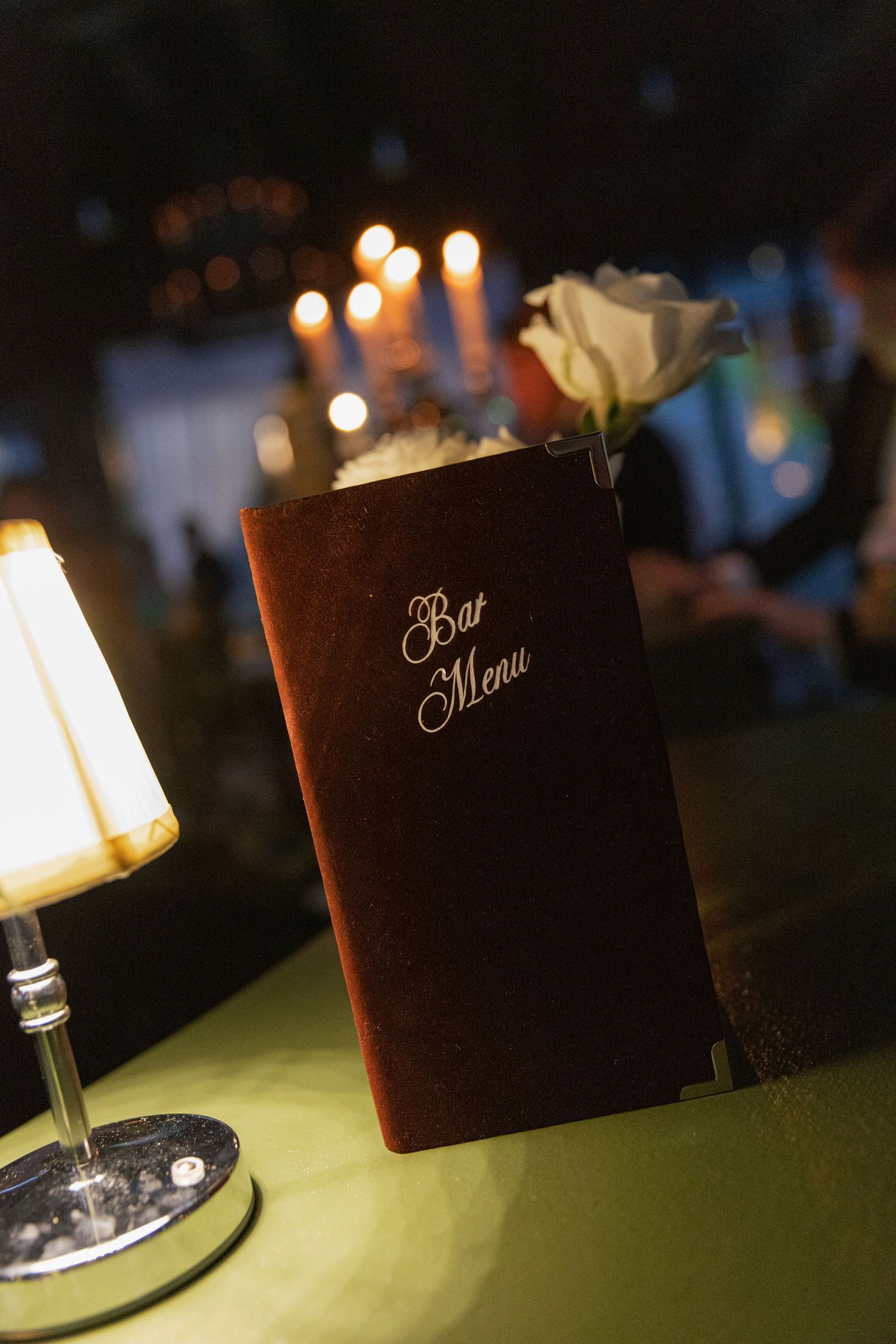 A velvet bar menu with a white rose sits on a green-covered bar beside a lamp, with glowing candles in the background.