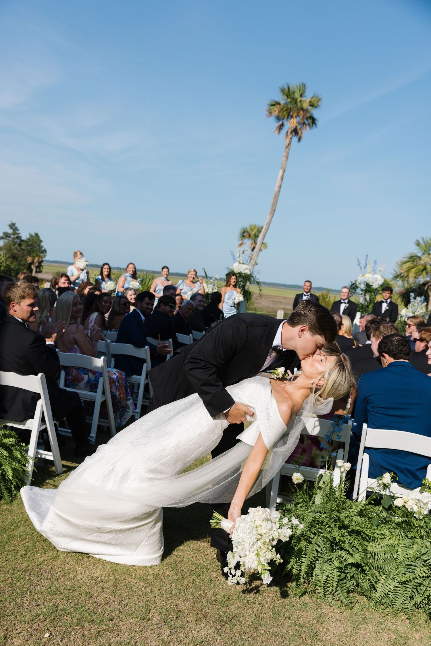 A romantic dip kiss between newlyweds during an outdoor wedding ceremony with palm trees and guests seated on white chairs.