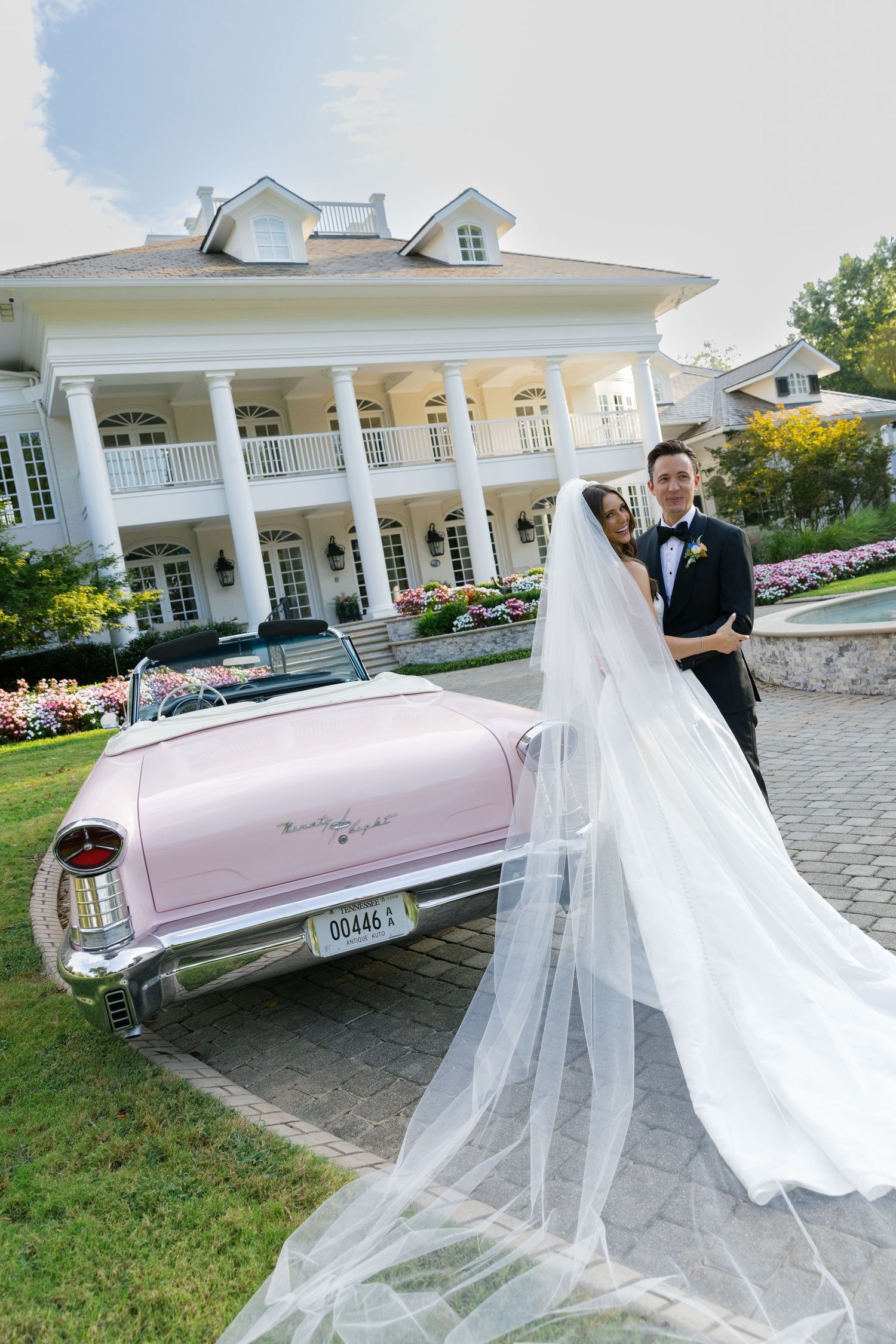 A wedding couple poses with a vintage pink Cadillac outside an elegant white mansion venue.