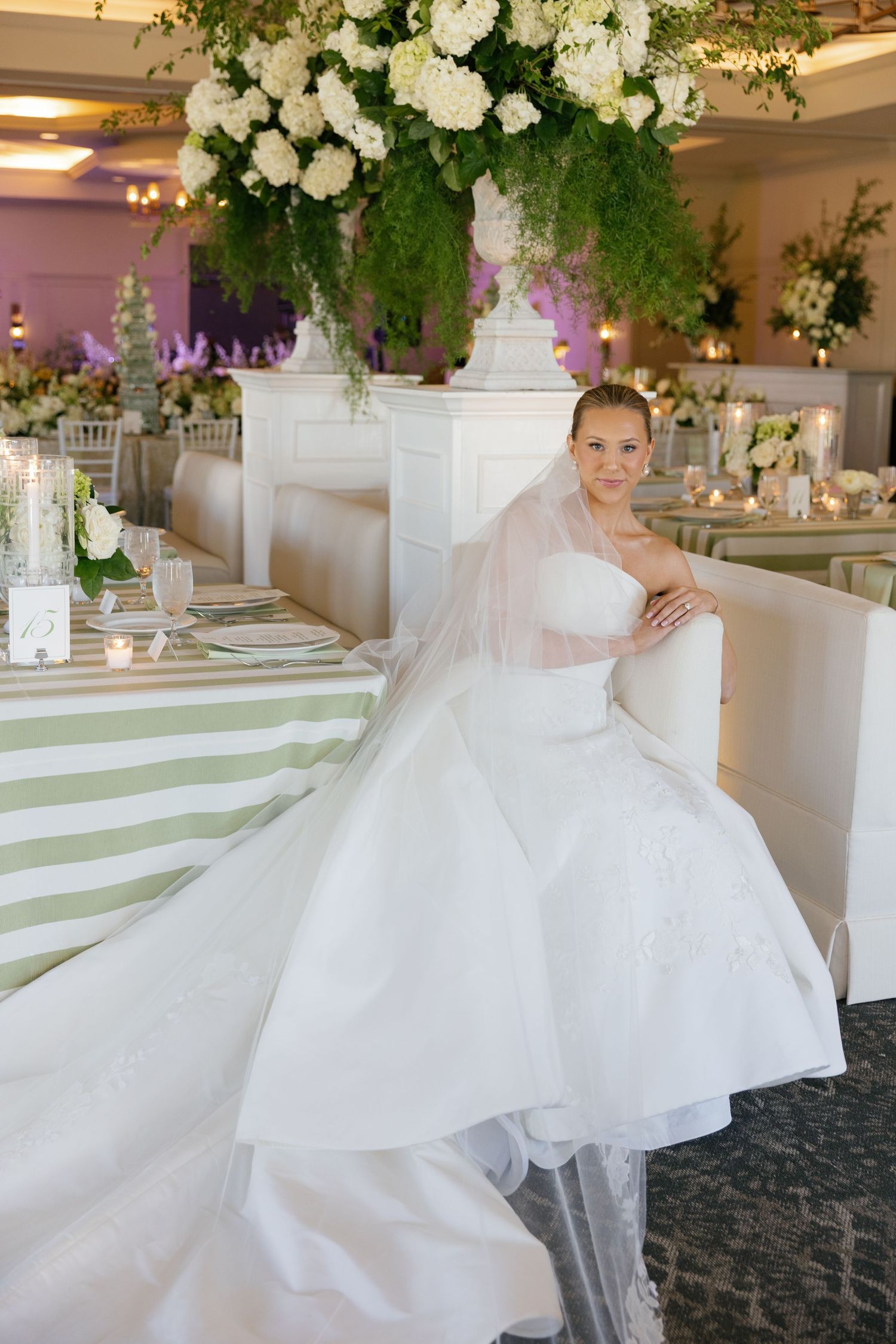 Bride in strapless white ballgown with veil sits beside elegant reception table with green stripe linens and lush floral arrangements.