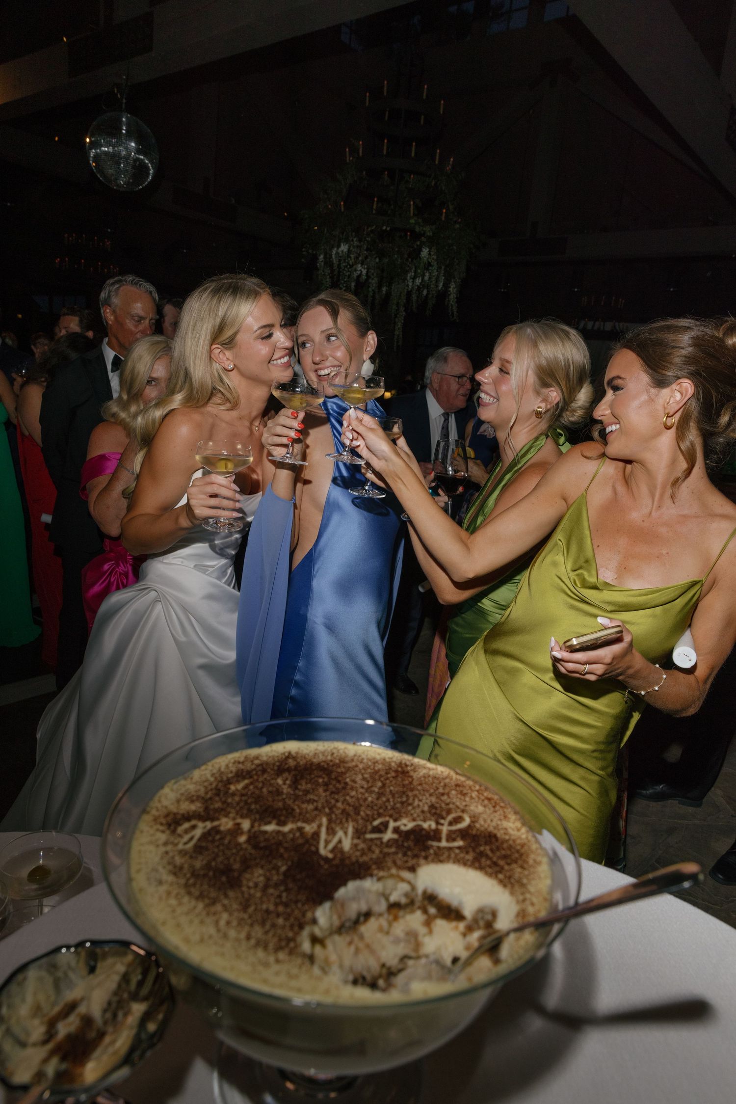 Bride and bridesmaids in blue and green satin dresses raise champagne glasses around a large tiramisu dessert at a lively wedding reception.