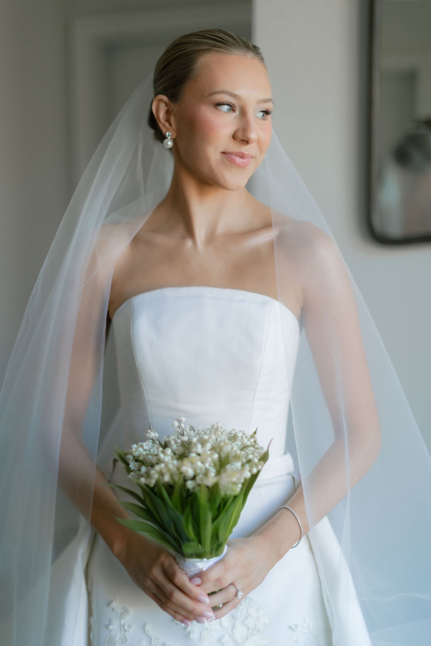 Elegant bride in strapless white gown and veil holding lily of the valley bouquet, gazing softly to the side.