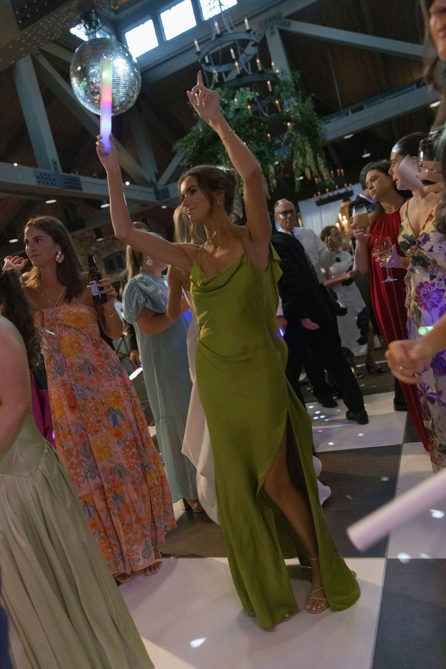 Guest in a green satin dress raises her arms and dances under a disco ball at a lively wedding reception with colorful dressed guests.