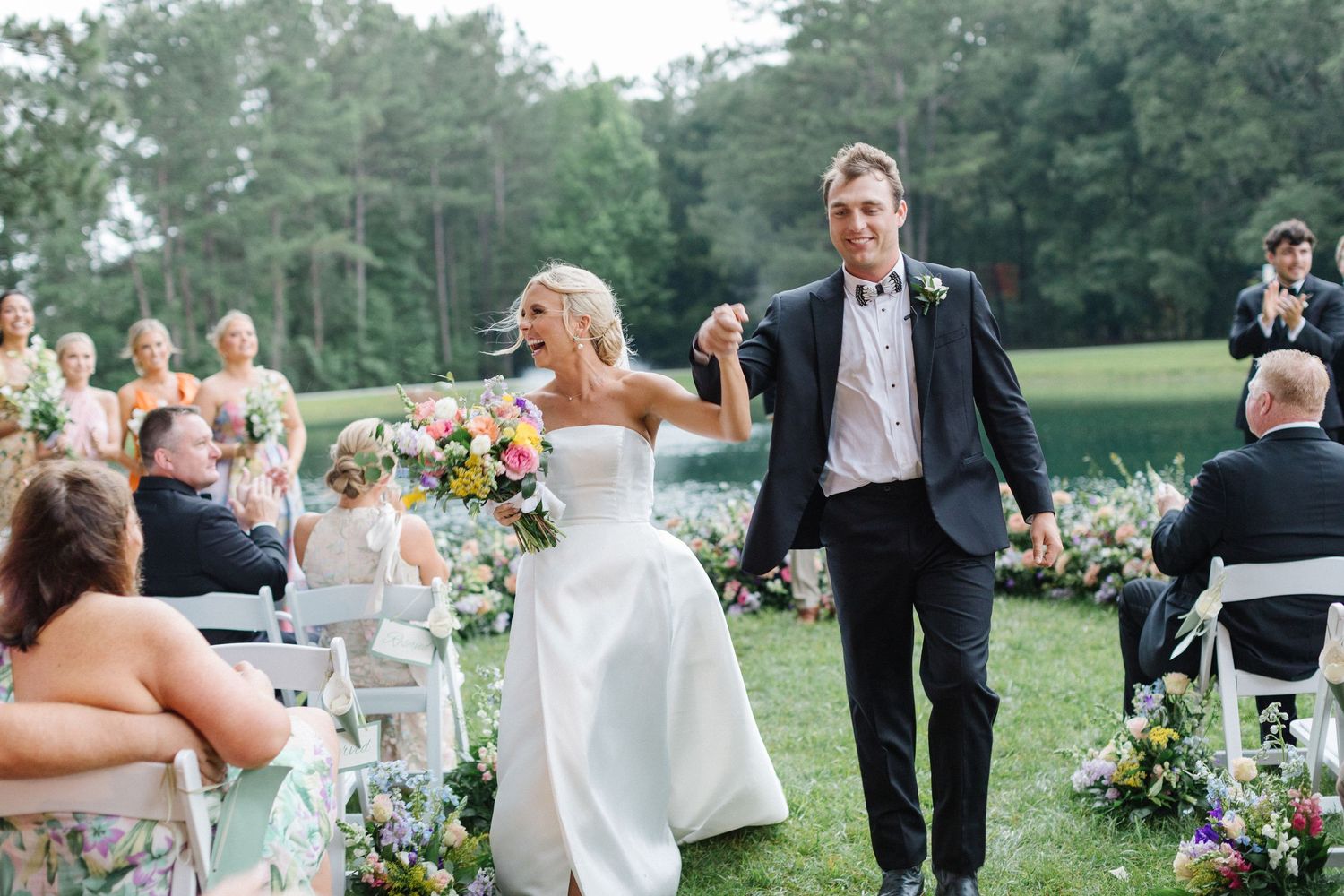 A joyful couple walks down the outdoor wedding aisle together as guests look on from white chairs on the lawn.