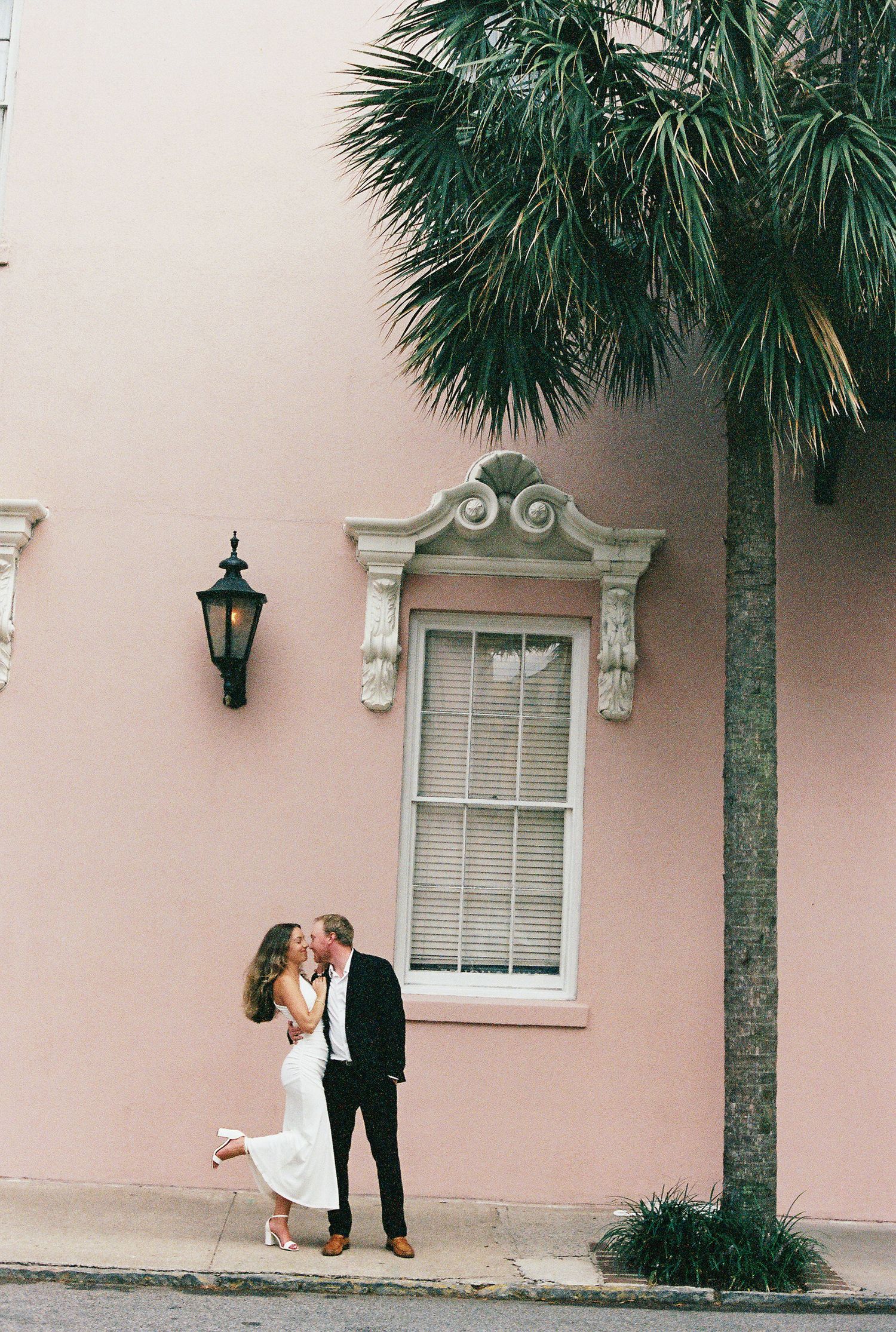 Romantic couple embraces against pink stucco wall with palm trees and vintage lanterns in Charleston's historic district.