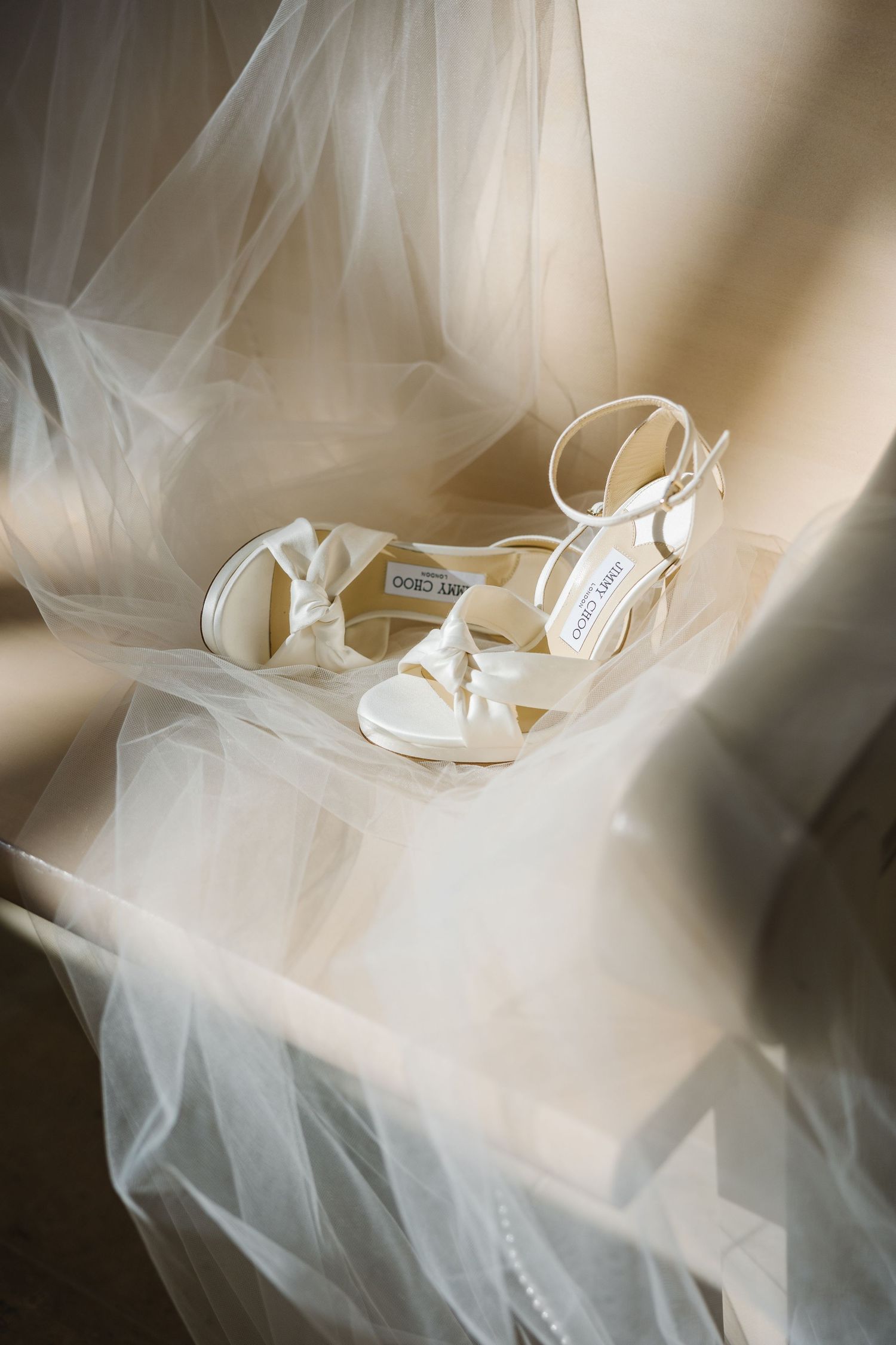 Elegant white bridal sandals with ankle straps placed on flowing tulle veil in soft natural lighting.