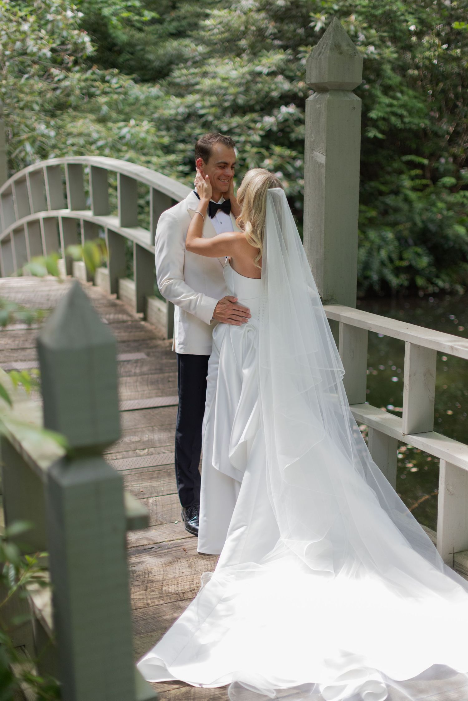 Bride in a strapless white gown with a long veil embraces her groom in a white dinner jacket on a wooden garden bridge.