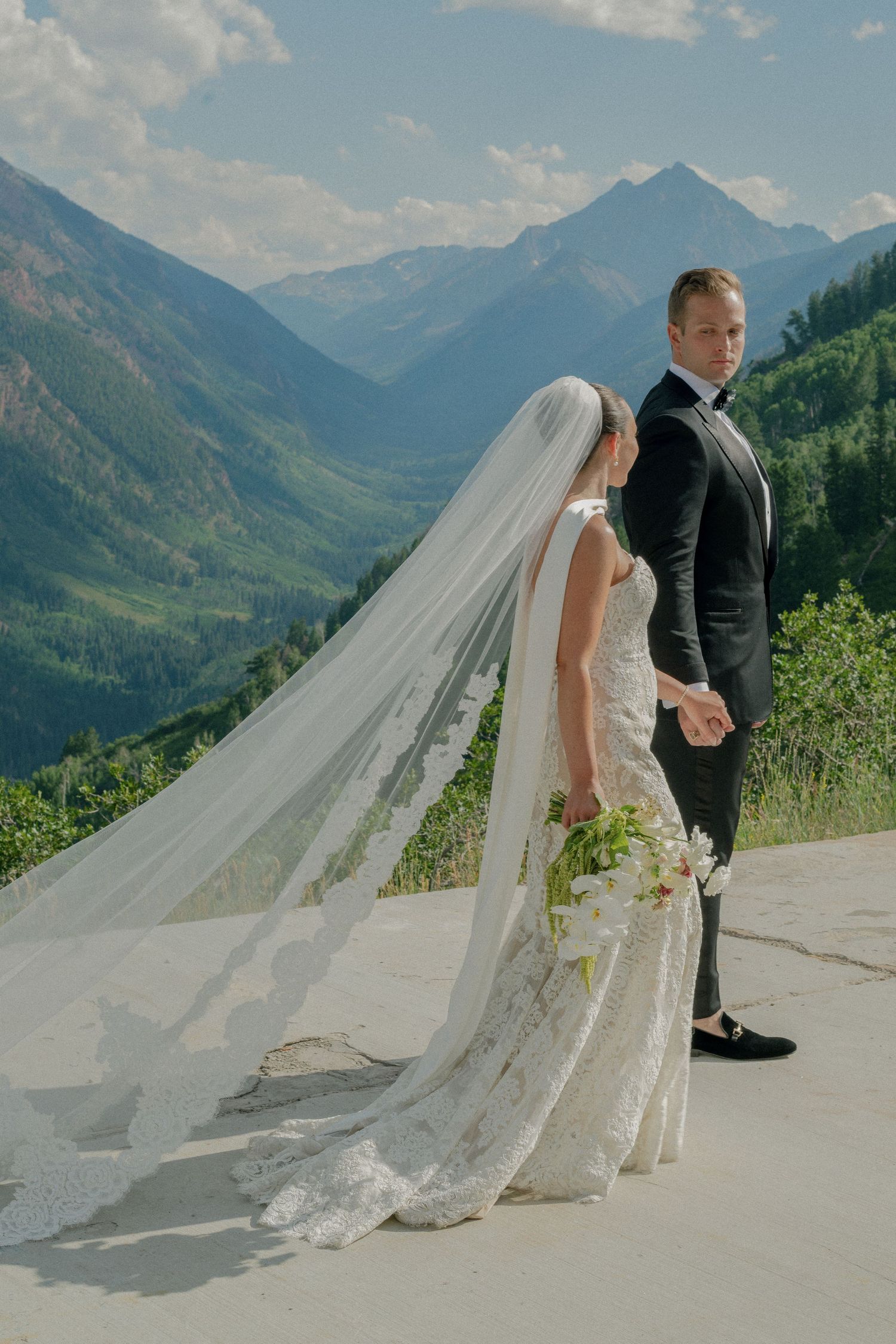A couple in wedding attire stands on a mountain overlook with dramatic valley views and peaks in the background.