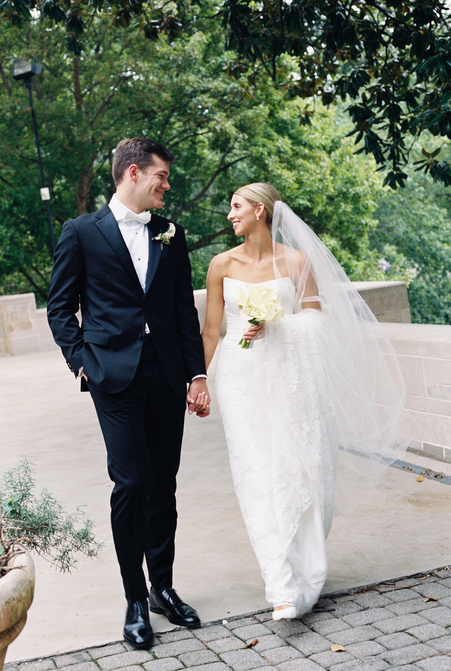 Newlyweds walk hand in hand along stone pathway surrounded by lush greenery wearing formal wedding attire.