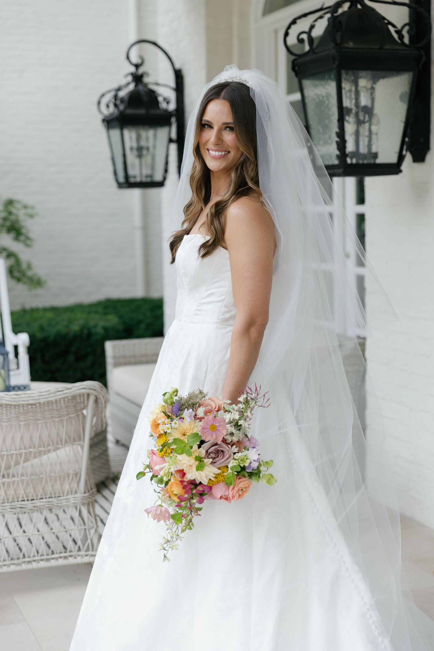 A bride in a white wedding gown holds a colorful bouquet of pink and peach flowers on a porch with lanterns.