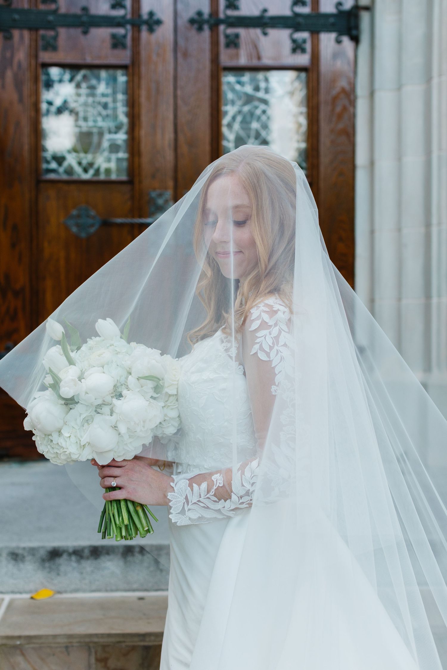 Elegant bride in lace gown and long veil holds white floral bouquet while standing by wooden church doors.