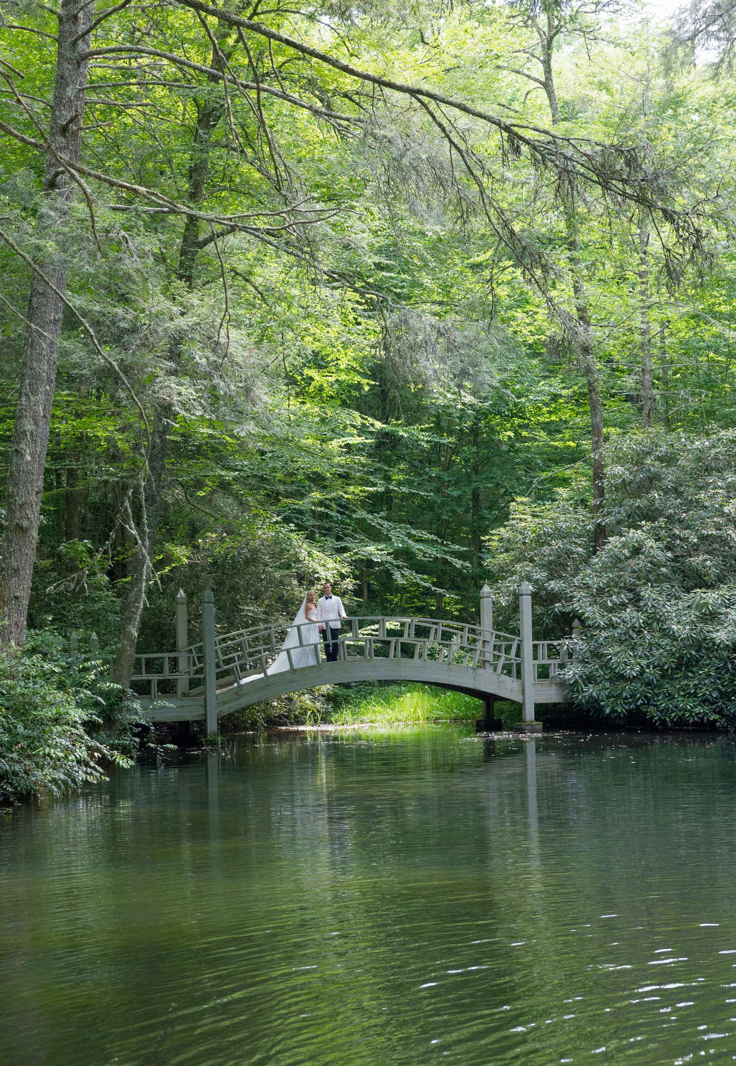 A bride and groom stand on a small arched bridge over a calm pond surrounded by lush green woodland.