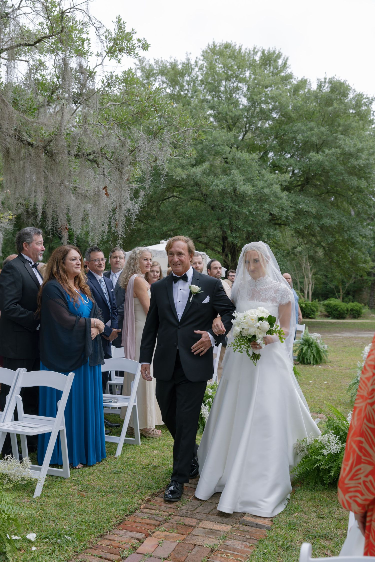 Father of the bride walks daughter down outdoor wedding aisle amid guests and Spanish moss-draped trees.