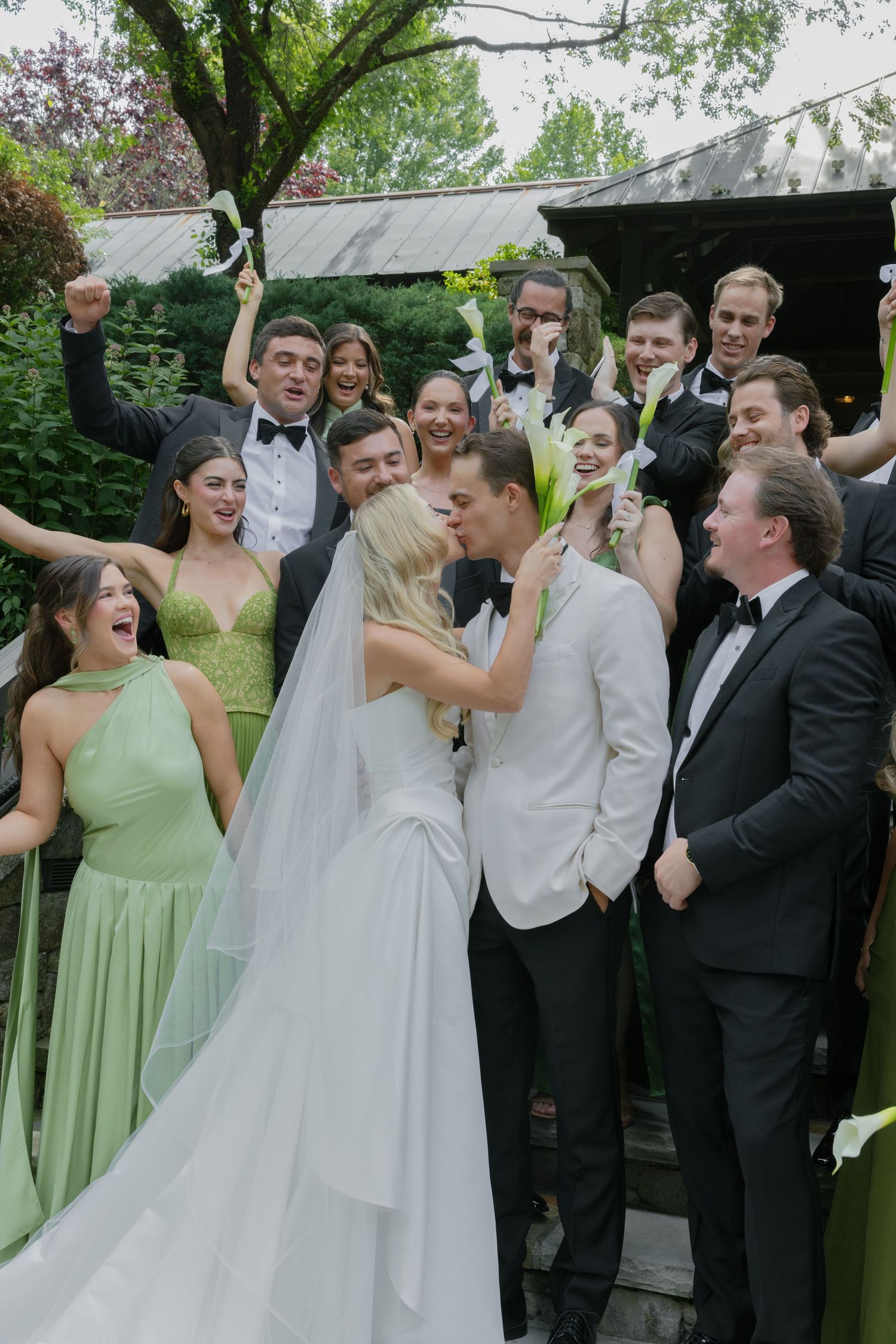 Bride and groom share a kiss on stone steps as cheering bridesmaids in green dresses and groomsmen in tuxedos celebrate around them.