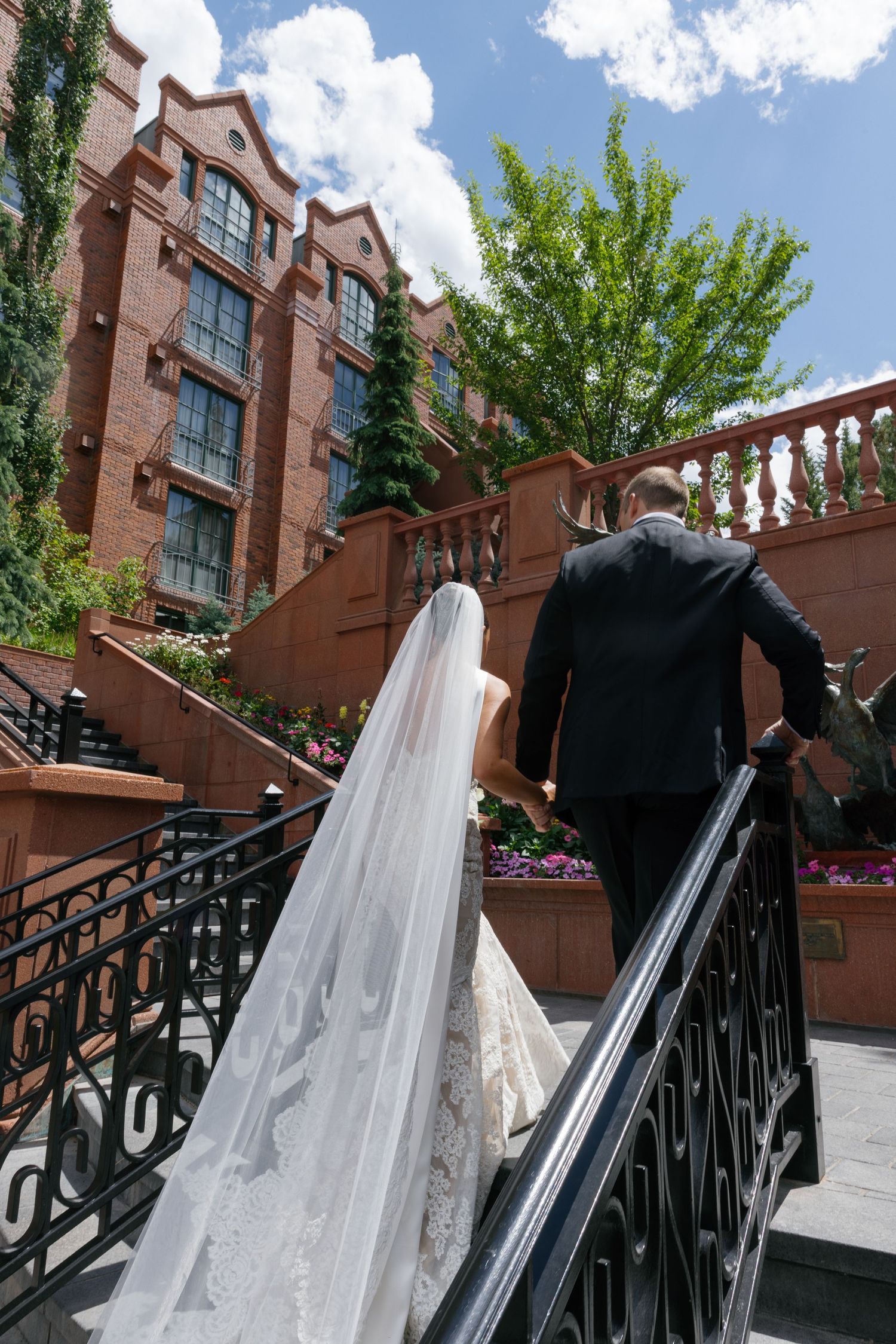 Wedding dresses and tuxedos displayed on black metal railings against a brick building exterior on a sunny day.