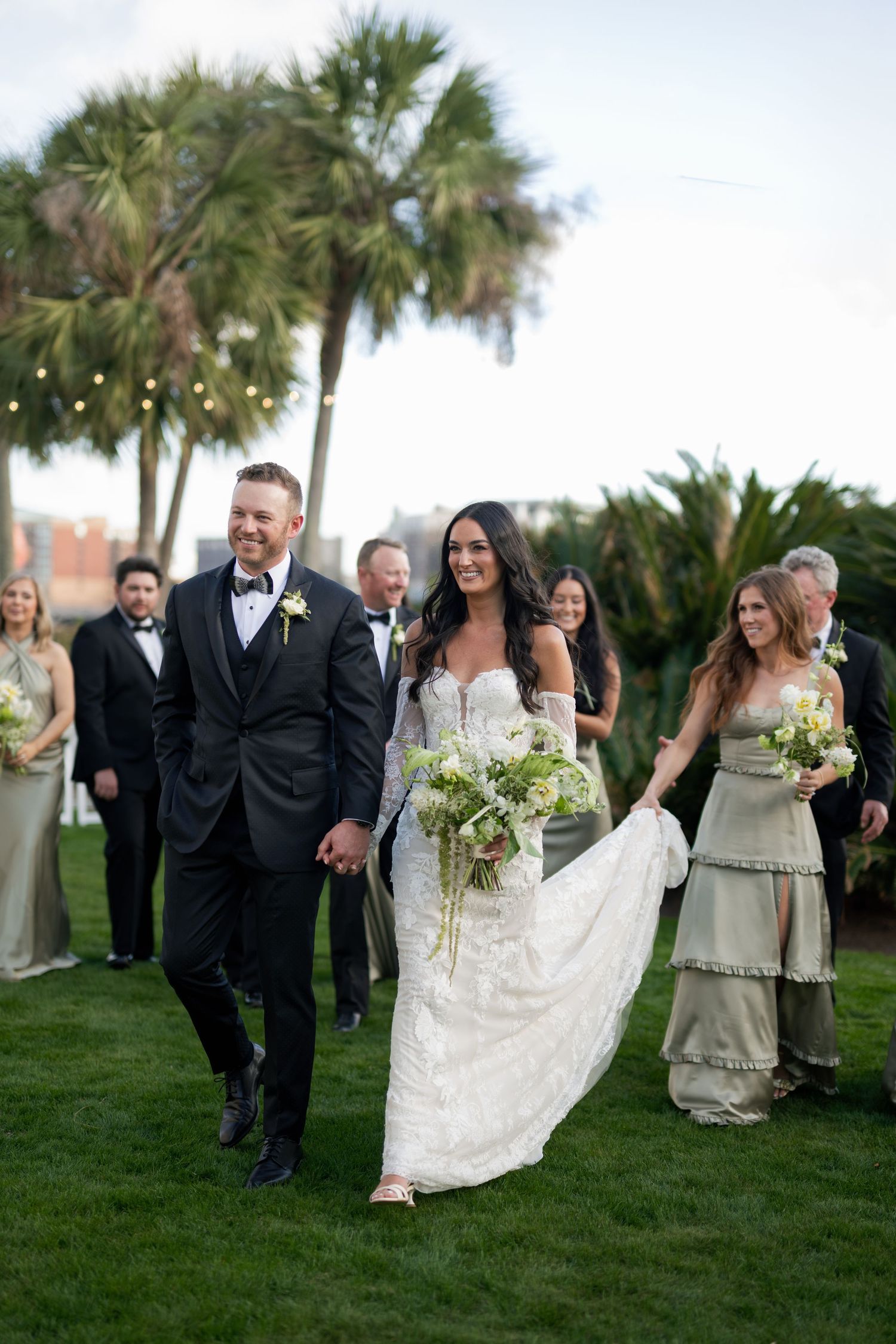 Bride and groom walk hand-in-hand after outdoor wedding ceremony, surrounded by wedding party near palm trees.
