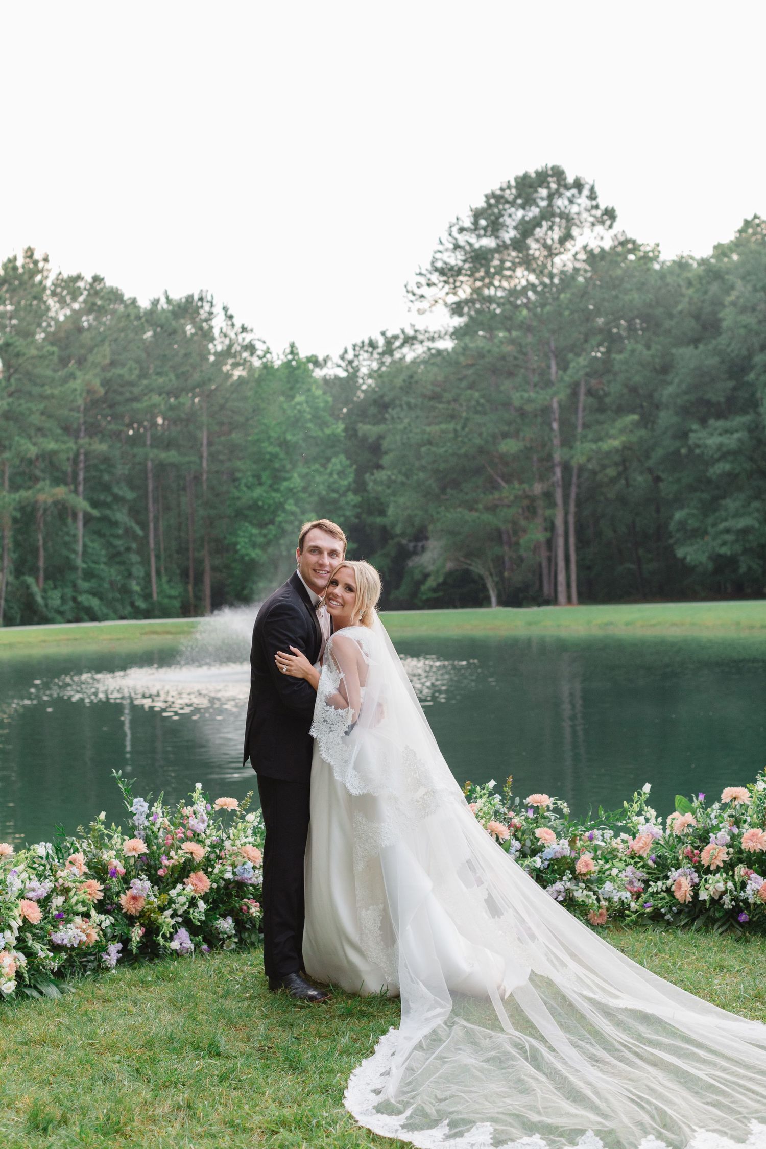 A romantic wedding couple shares an intimate moment by a serene lake surrounded by pine trees and colorful flowers.