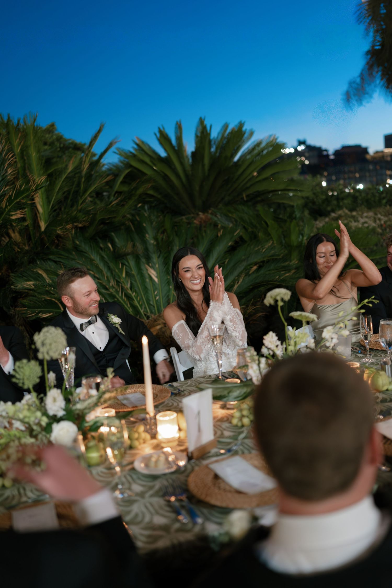 Bride and guests clapping and laughing at an elegant outdoor wedding reception dinner at dusk with candles.