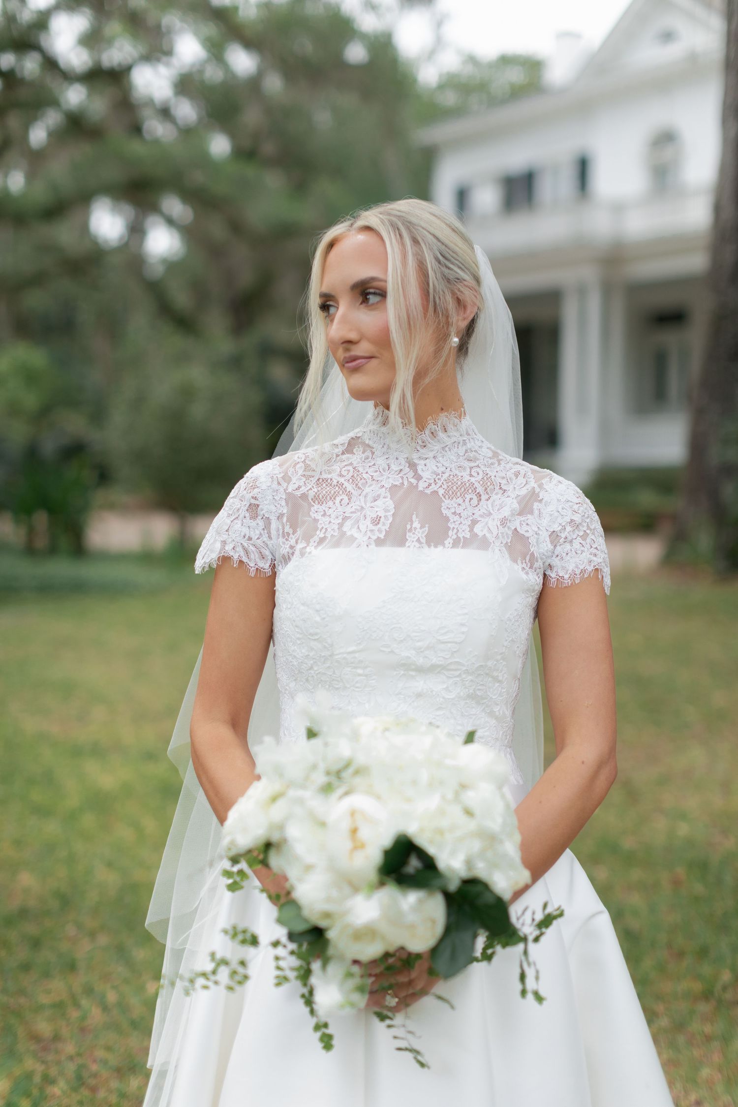 Blonde bride in lace high-neck wedding gown holding white peony bouquet standing on green lawn before white mansion.