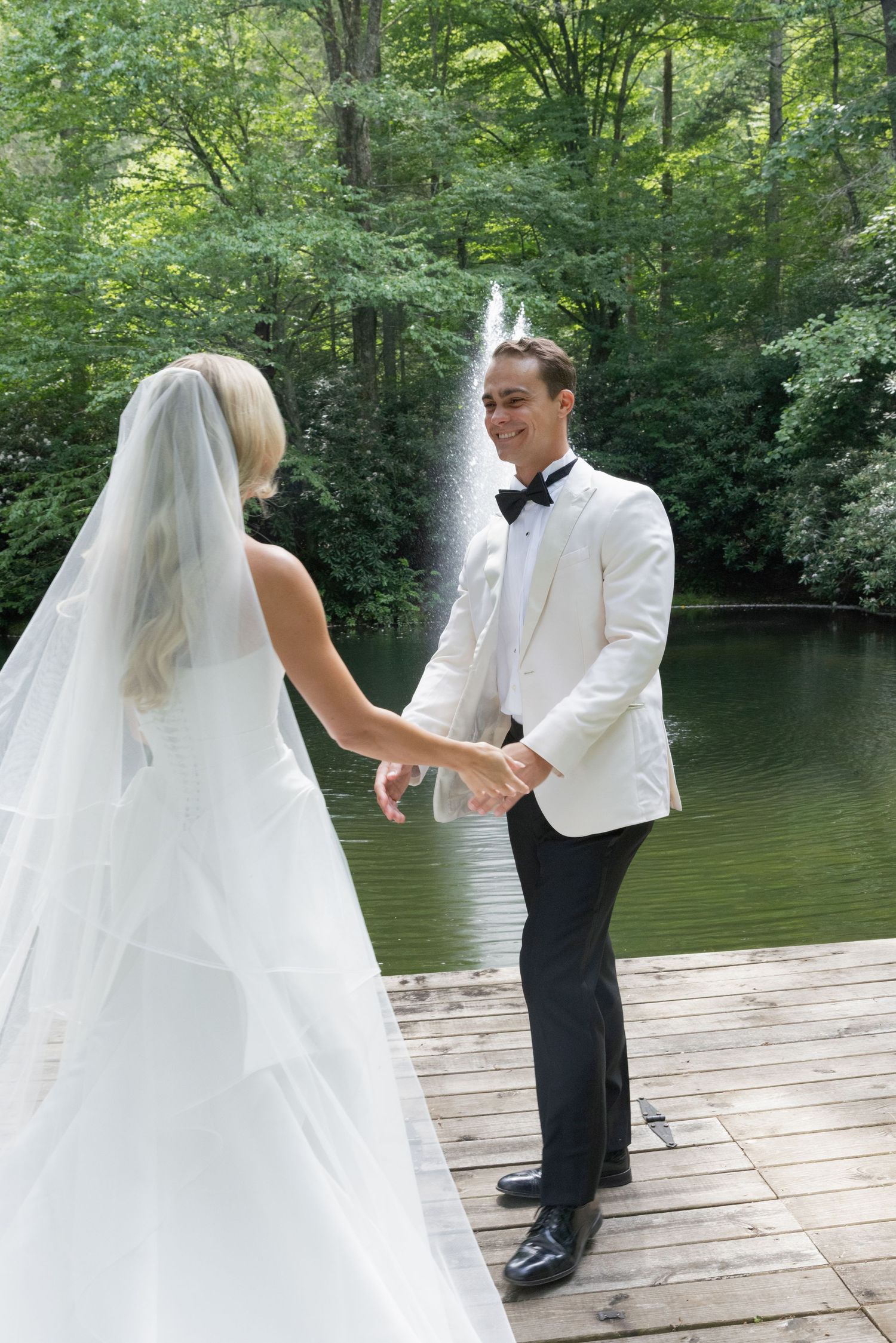 Groom in white tuxedo jacket and bow tie holds hands with a veiled bride in a wedding gown on a wooden dock by a lake and fountain.