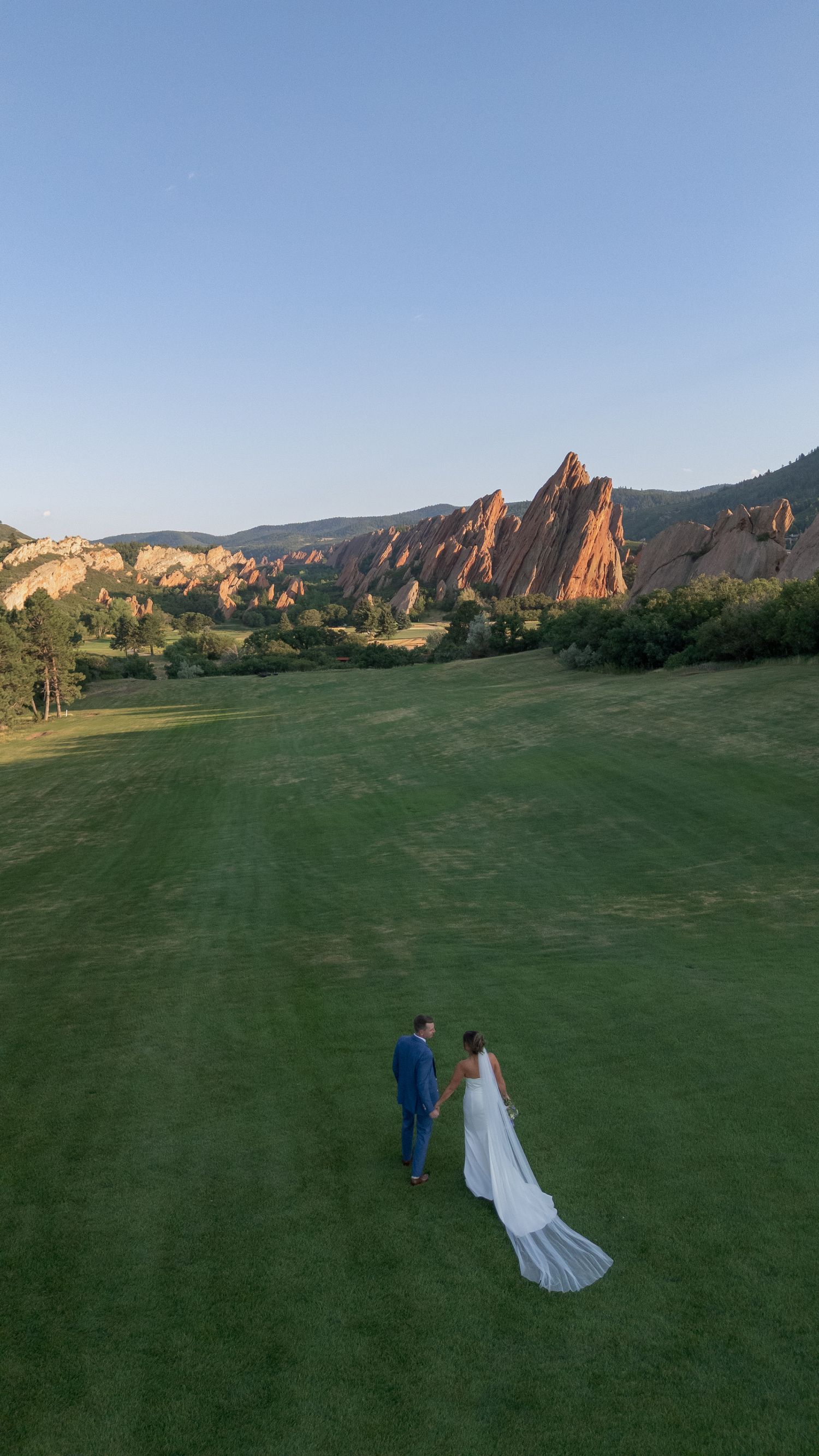 Bride and groom walk hand in hand across a green lawn toward red sandstone rock formations at sunset in a scenic Colorado wedding venue.