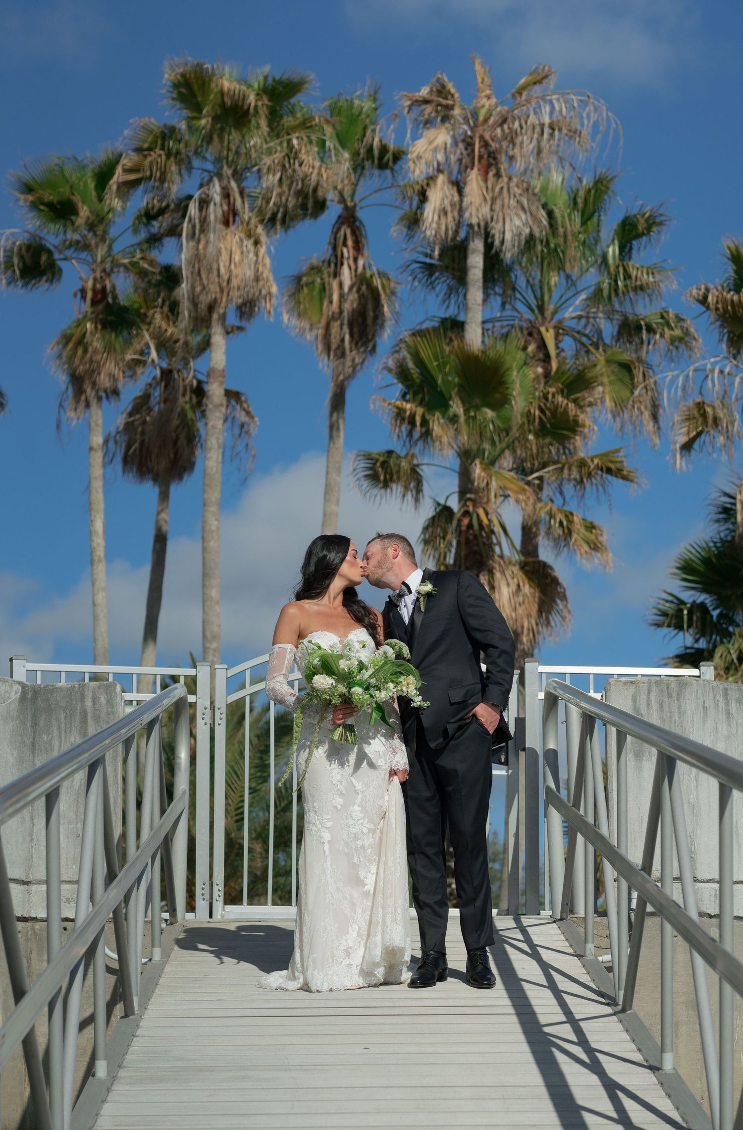 A romantic wedding couple stands on a wooden boardwalk surrounded by tall palm trees against a bright blue sky.