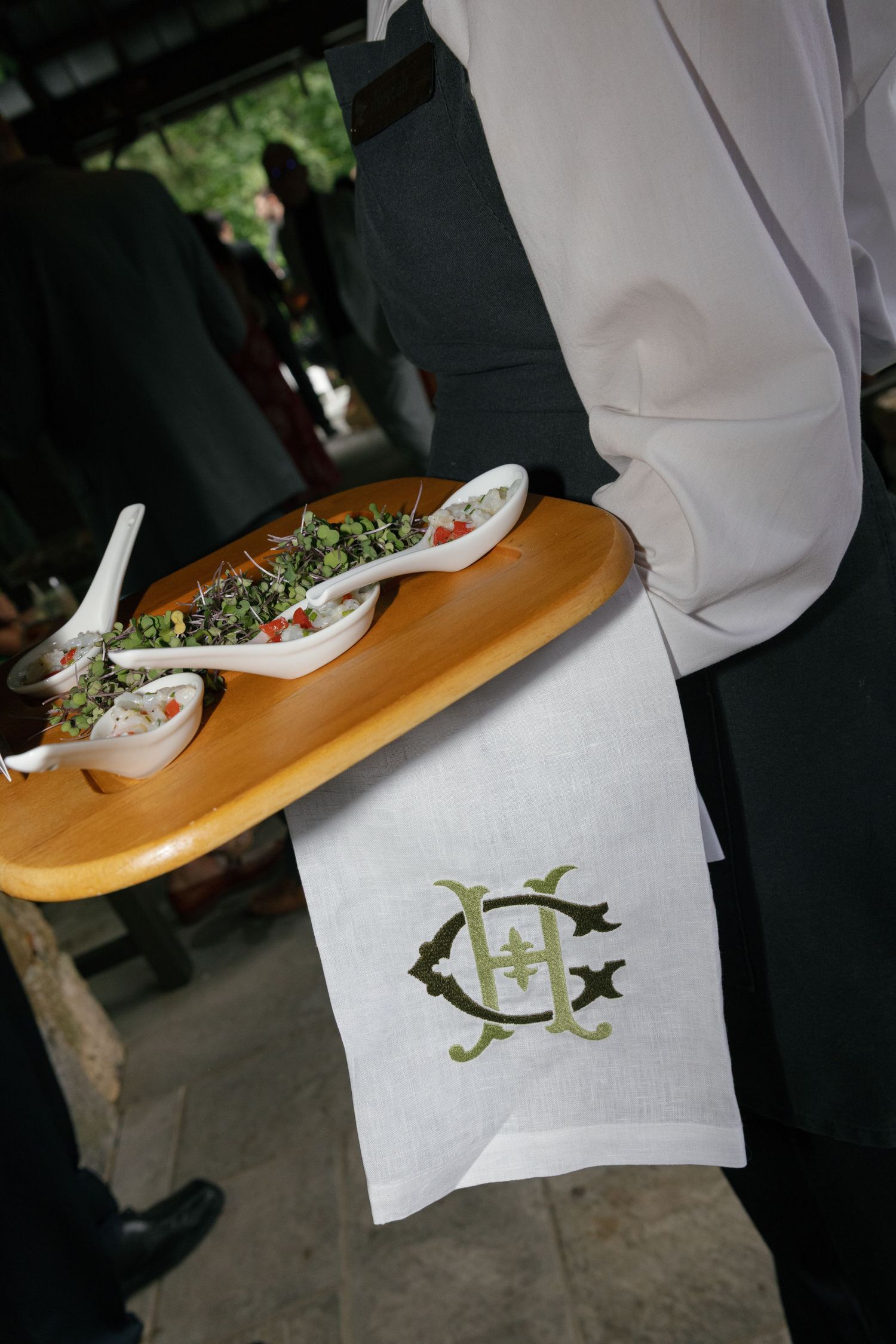 Server holding a wooden tray of appetizers in white spoons, with a monogrammed linen napkin draped over the arm at an outdoor event.