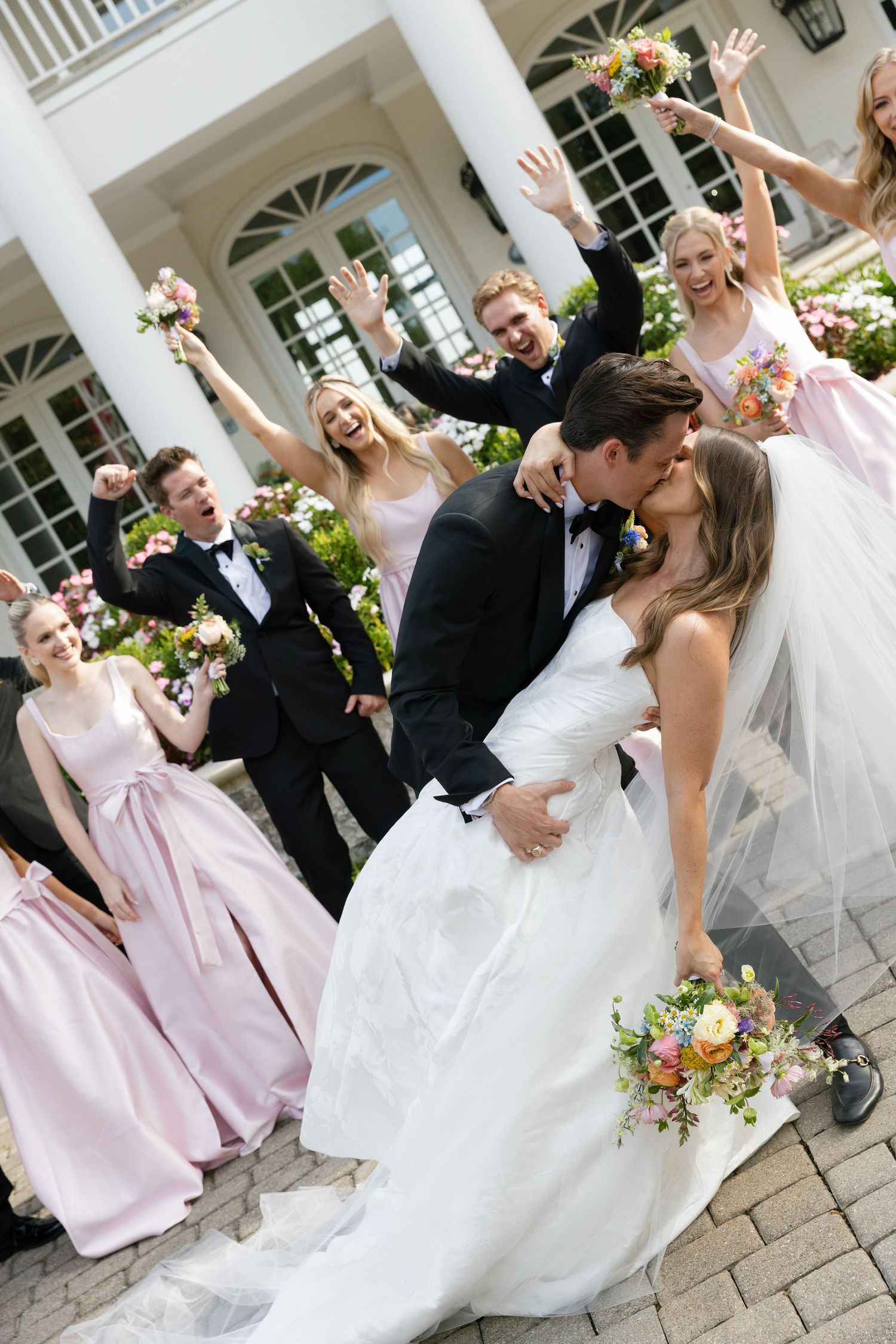 Wedding party celebrates joyfully on stone patio outside elegant white building with arched windows.