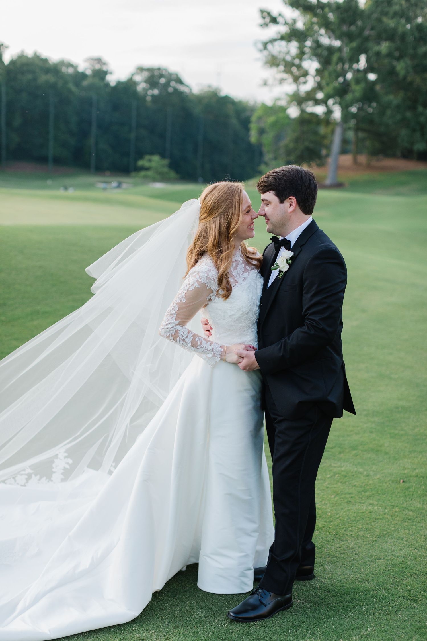 Wedding couple embraces on a lush green lawn with flowing white veil catching the breeze.