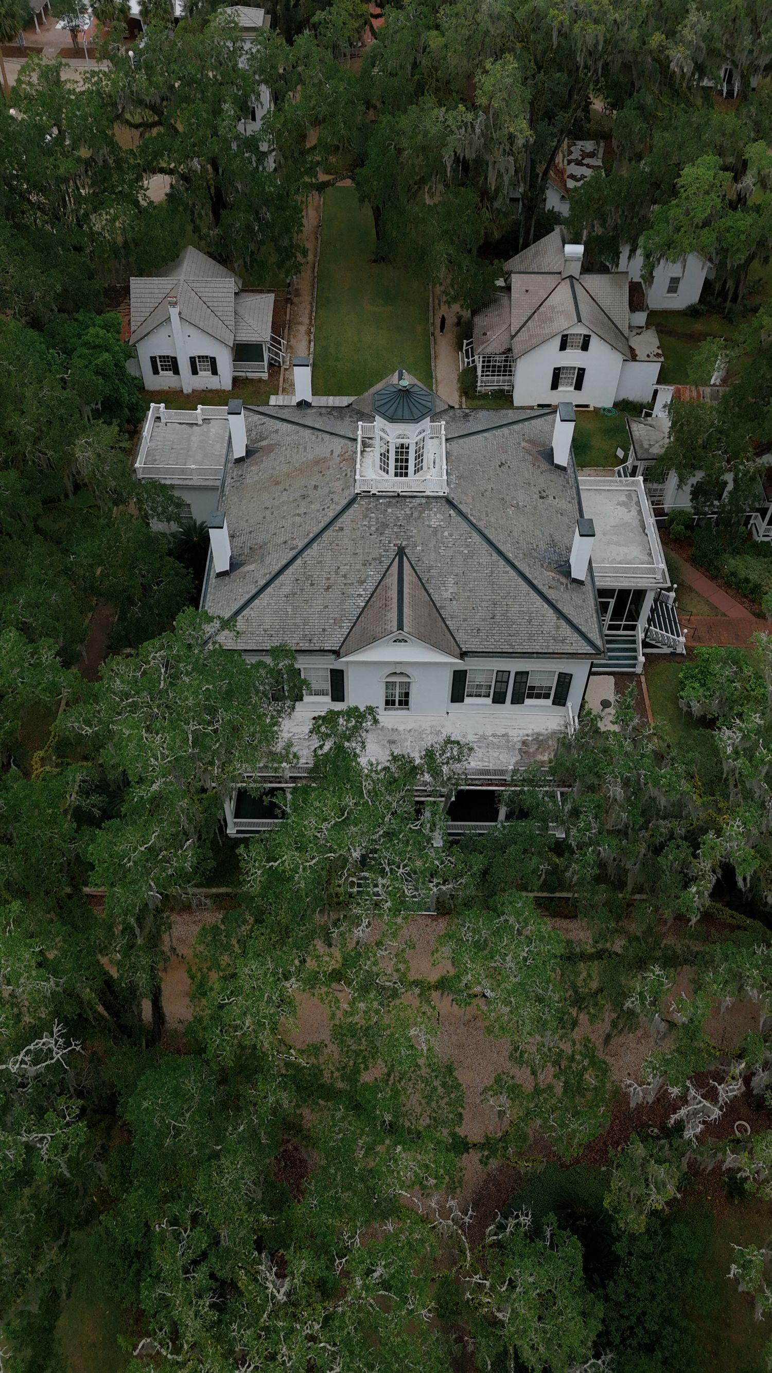 Aerial view of a historic Southern plantation home surrounded by moss-draped trees, featuring a cupola and white outbuildings.