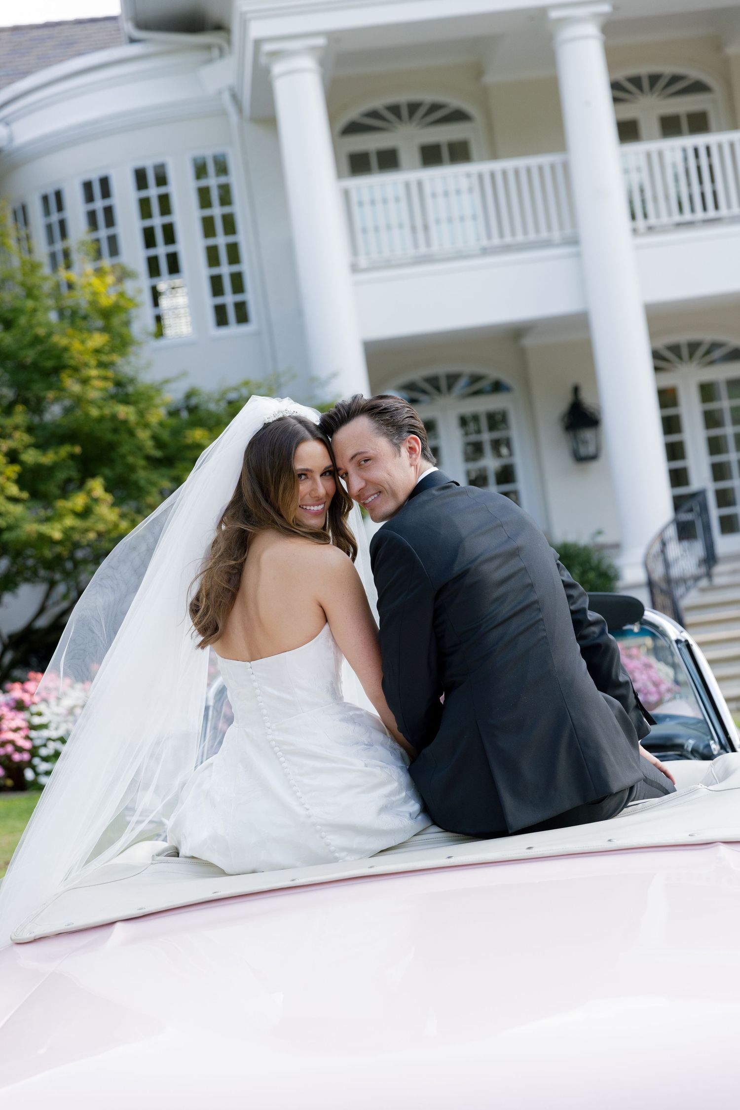 Newlyweds share an intimate moment while seated in a classic pink car outside a grand white columned mansion.
