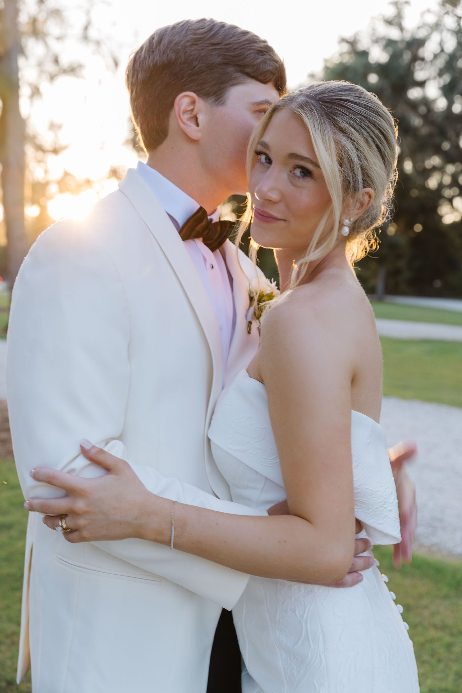 A couple shares an intimate moment at sunset in a park while wearing elegant white wedding attire.