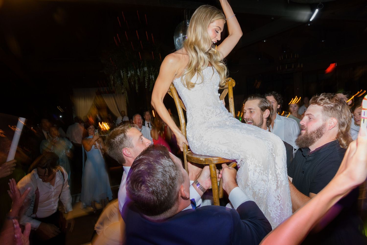 Bride in a white lace wedding gown is lifted on a chair by cheering guests during a lively reception dance.