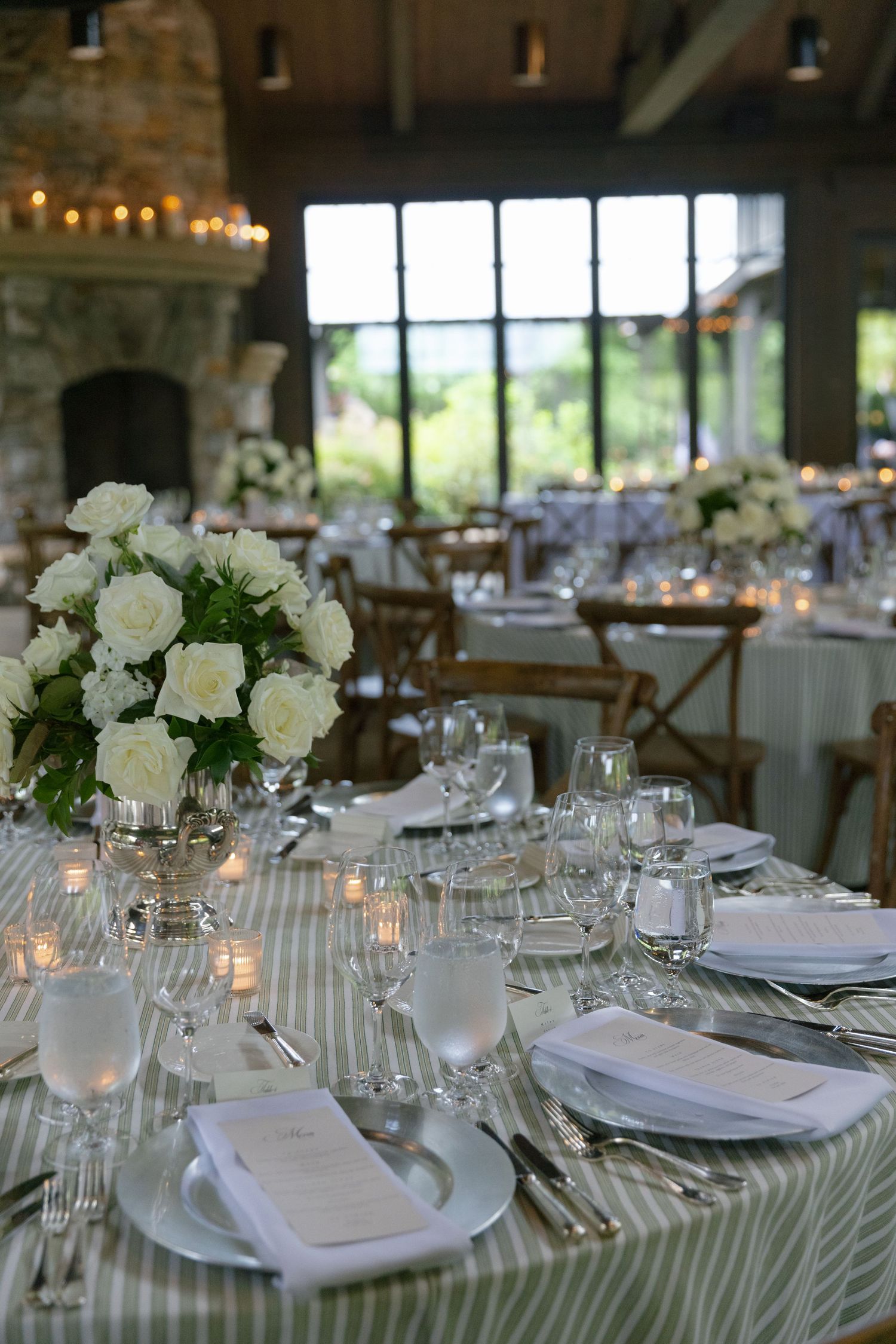 An elegant wedding reception table with striped green linens, white rose centerpieces, candles and place settings beside large windows.