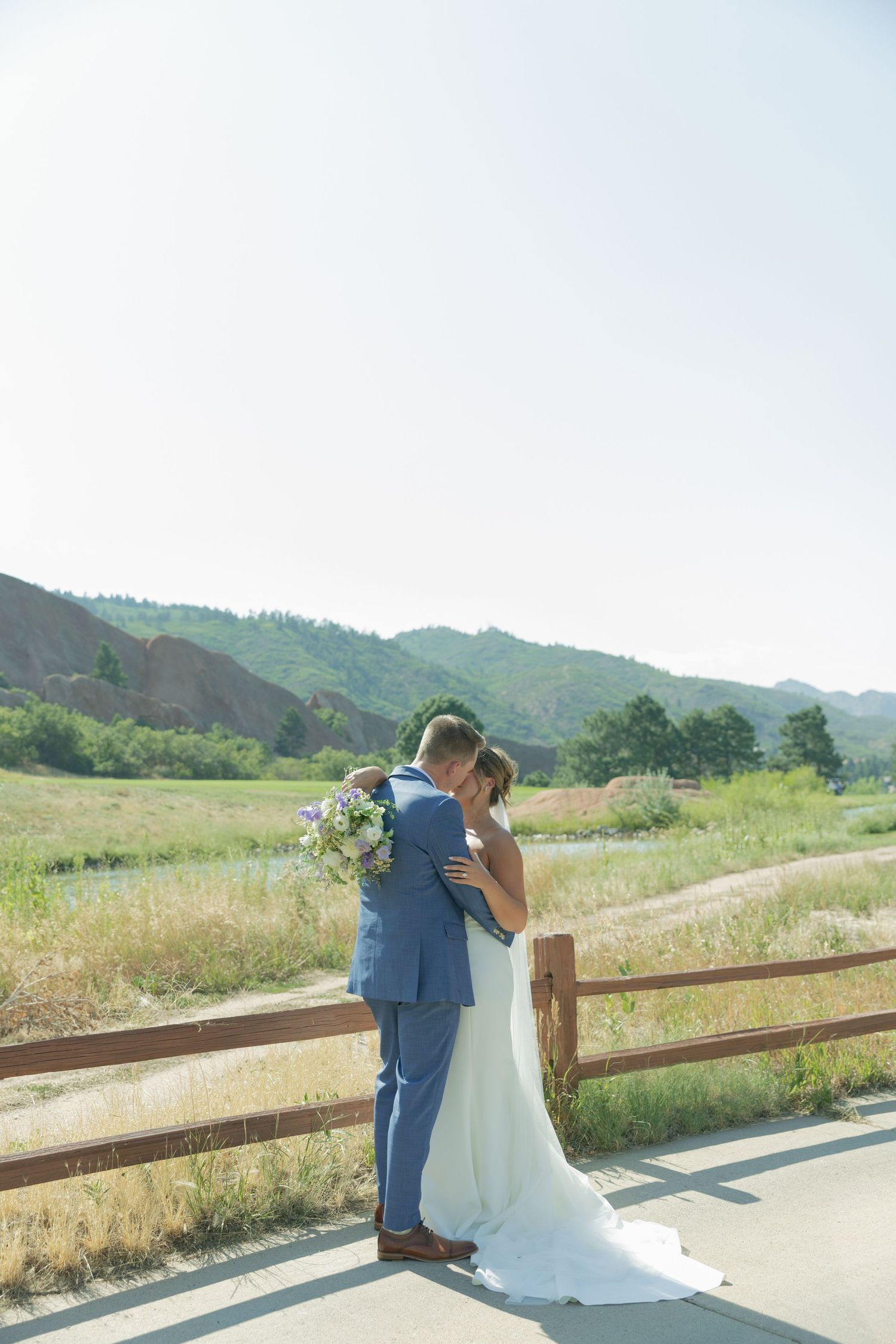 Bride in a white gown and groom in a blue suit embrace on a paved path by a wooden fence, with green hills in the background.