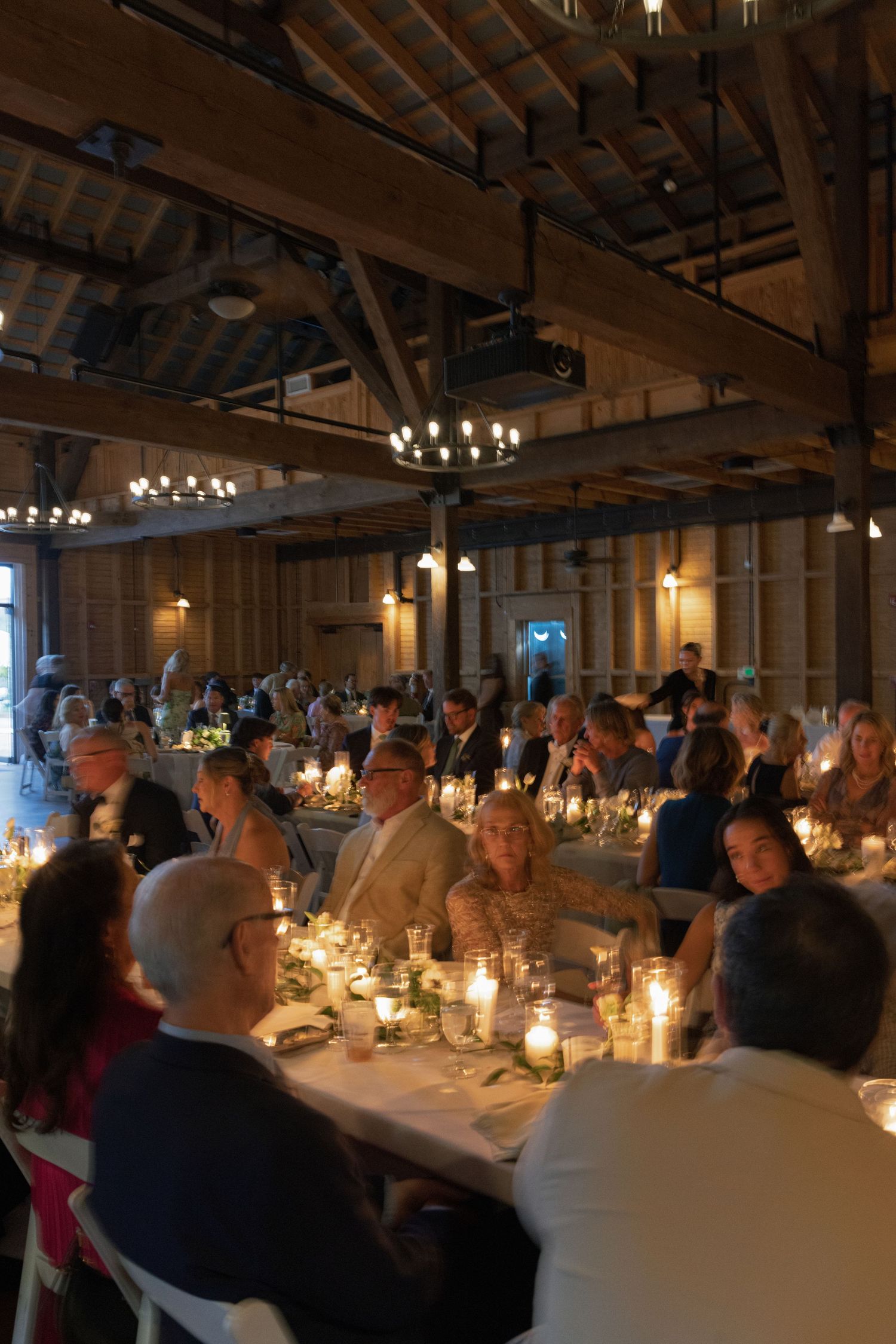 Wedding reception guests dine by candlelight inside a warmly lit rustic barn venue with exposed wooden beam ceiling.
