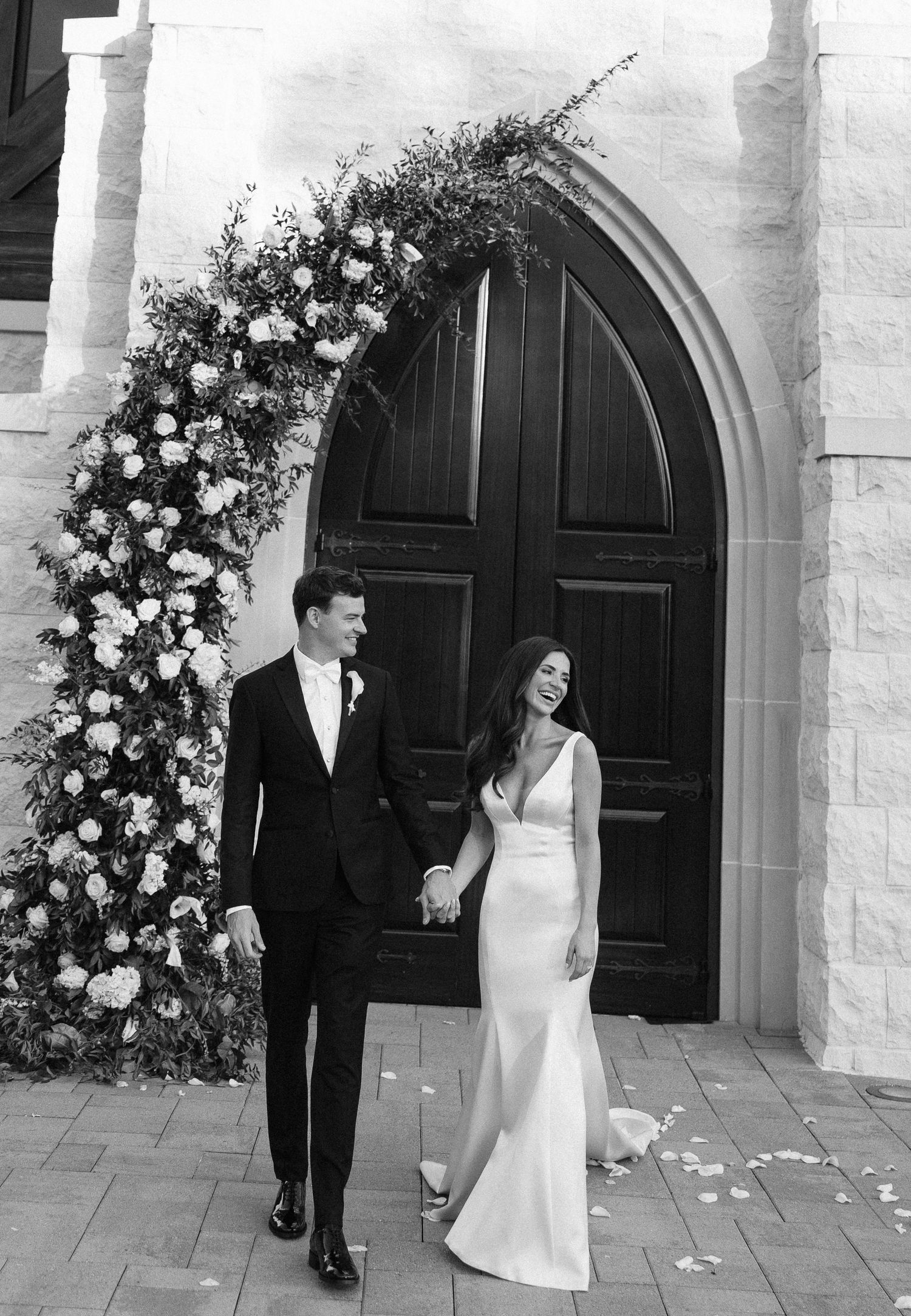 A newlywed couple walking outside a grand church archway adorned with elegant white floral arrangements in black and white.