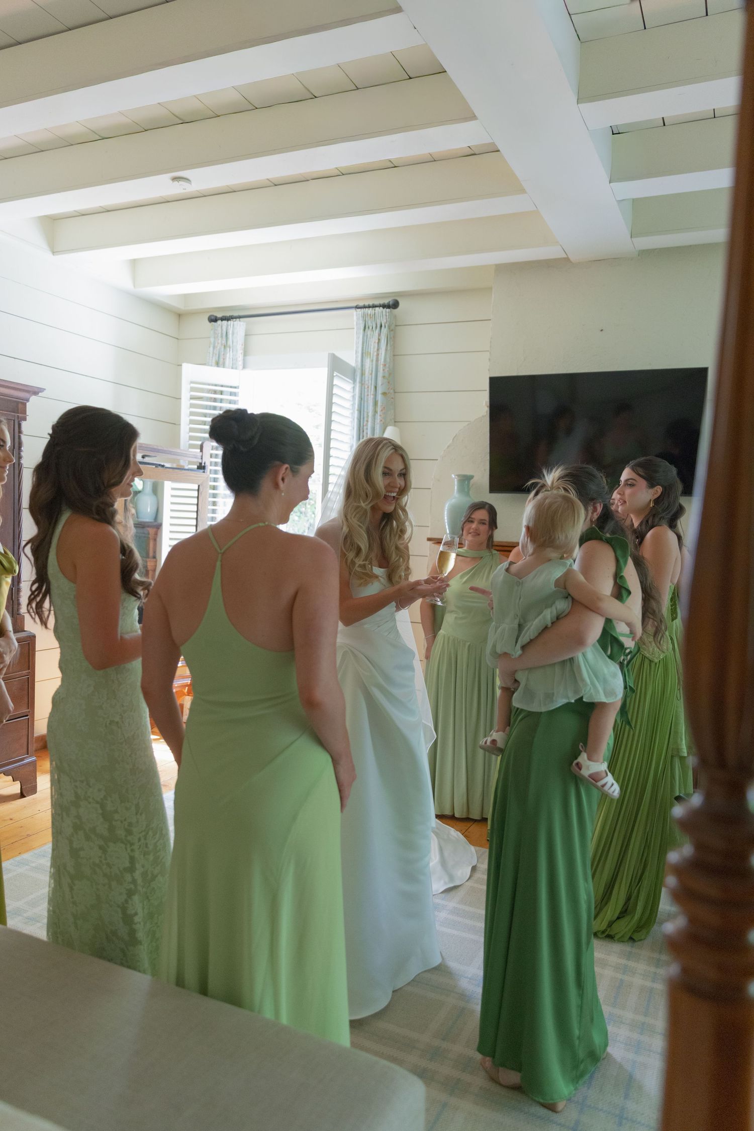 A bride in a white gown chats with bridesmaids in green dresses, one holding a toddler, in a bright room with white beamed ceilings.