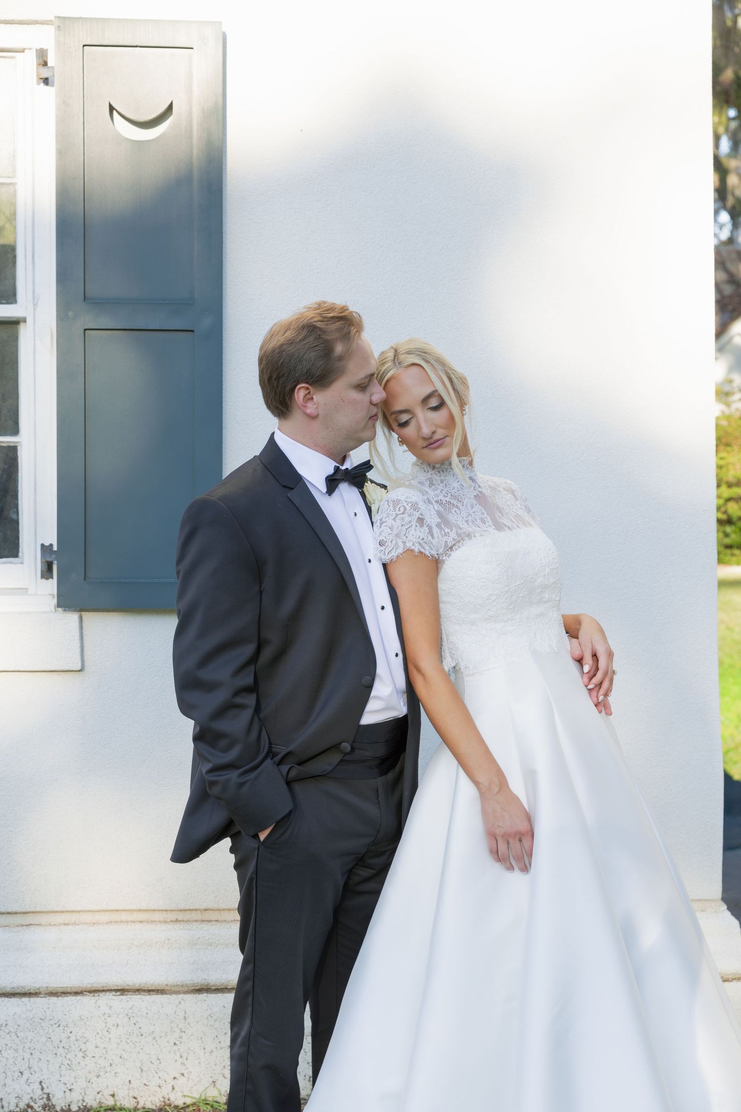 Bride in white lace gown leans on groom in black tuxedo outside white building with blue shutters.