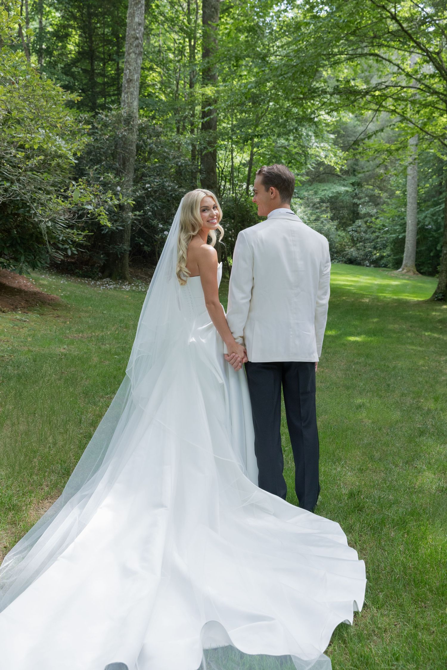 A bride in a flowing white gown with a long veil holds hands with her groom in a white jacket on a sunlit green lawn.